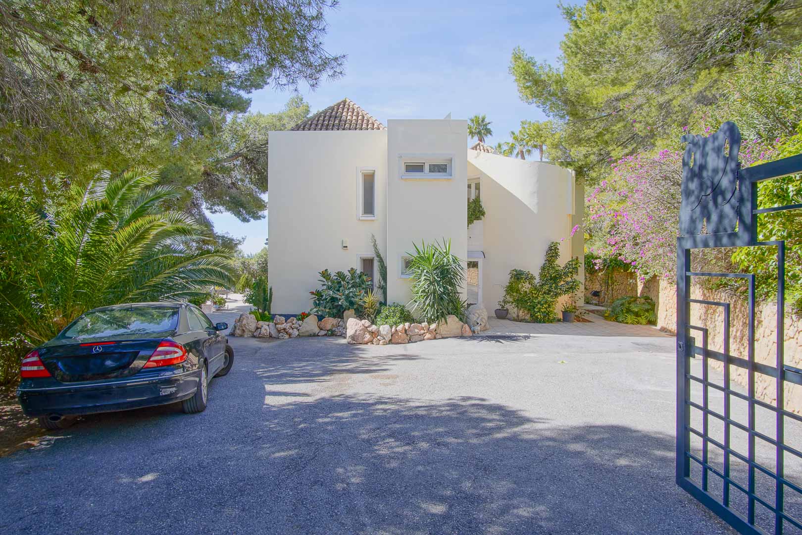 Hauptfassade der Villa im mediterranen Stil mit großzügigem gepflastertem Parkplatz und sicherem Eingang, umgeben von Pinien und üppiger Vegetation.