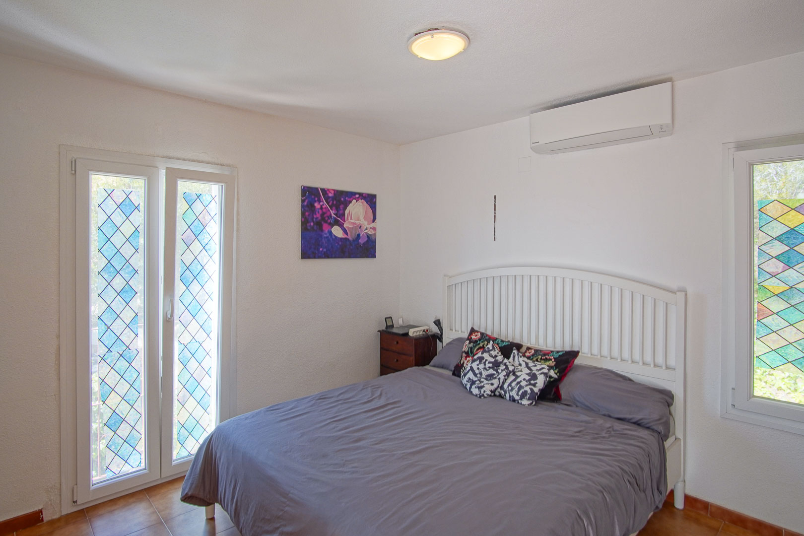 Helles Schlafzimmer mit Klimaanlage, weißem Kopfteil und Fenstern mit dekorativem, rautenförmigem Buntglas-Design.