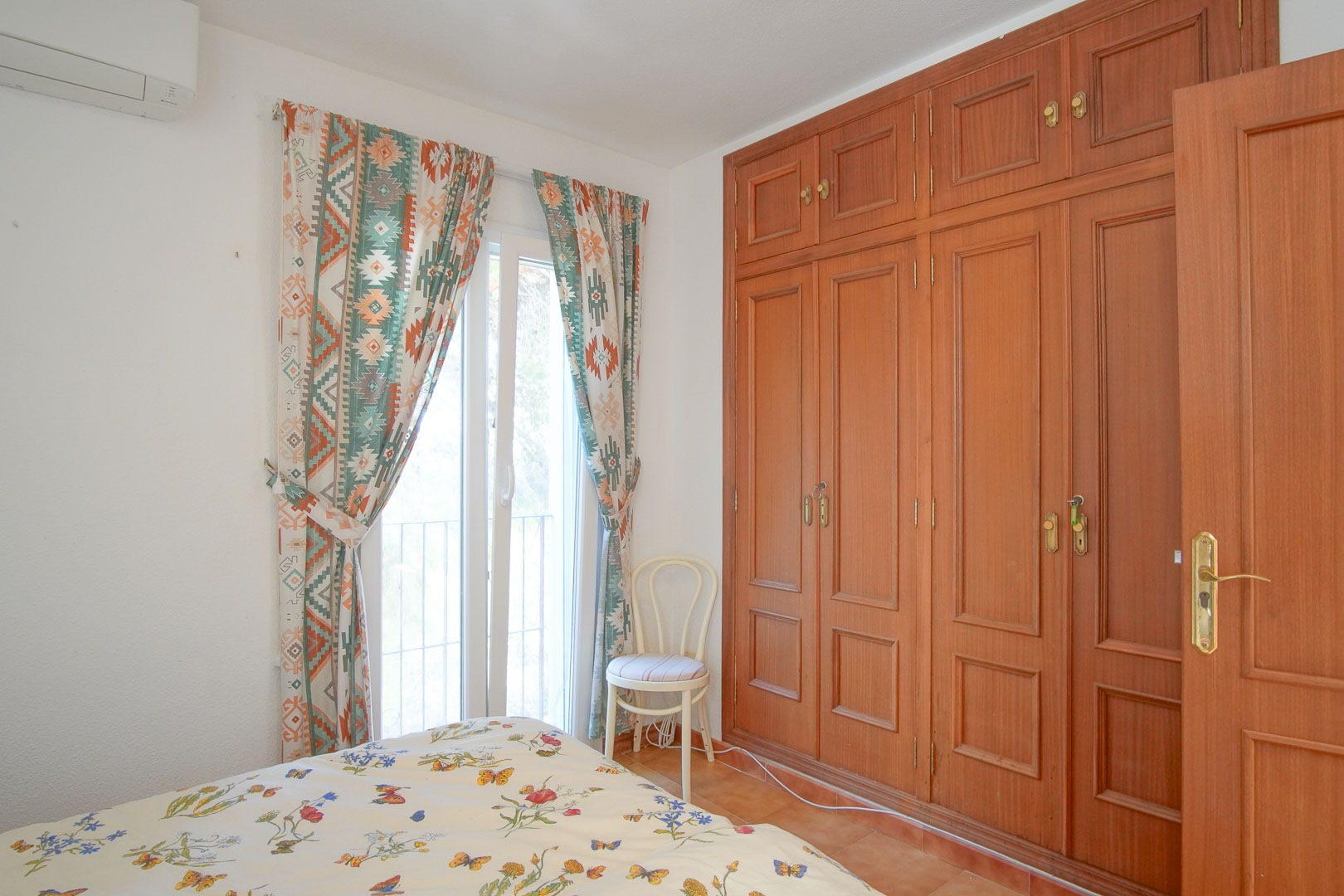 Helles Schlafzimmer mit raumhohen Einbauschränken aus Holz, Klimaanlage und direktem Zugang nach draußen durch eine Fenstertür mit gemusterten Vorhängen.