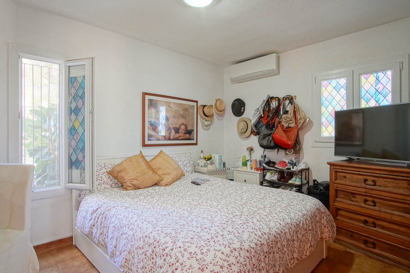 Helles Schlafzimmer mit Terrakotta-Fliesen, Split-Klimaanlage und dekorativen Buntglasfenstern. Ein klassisches Holzschränkchen ergänzt den lichtdurchfluteten Raum.