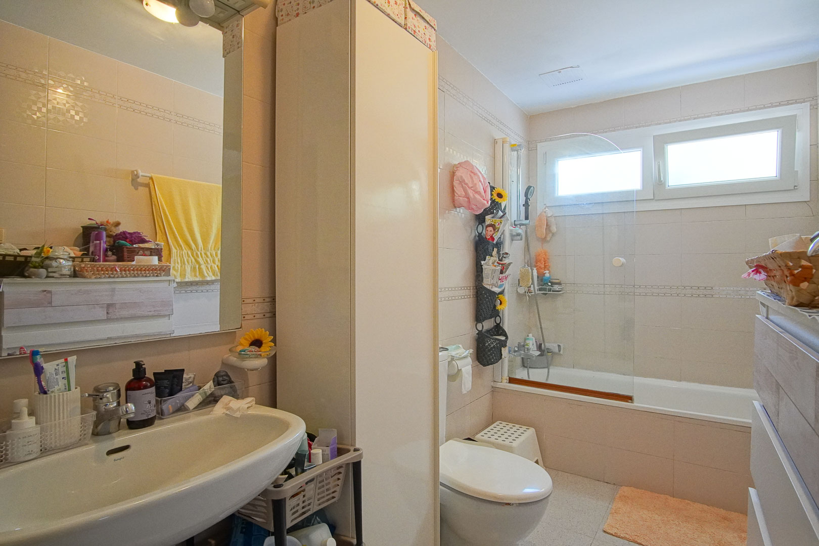 Komplettes Badezimmer mit Badewanne, Glastrennwand, weißem Waschbecken und Hochschrank. Beige Wandfliesen und ein Fenster bieten natürliches Licht.