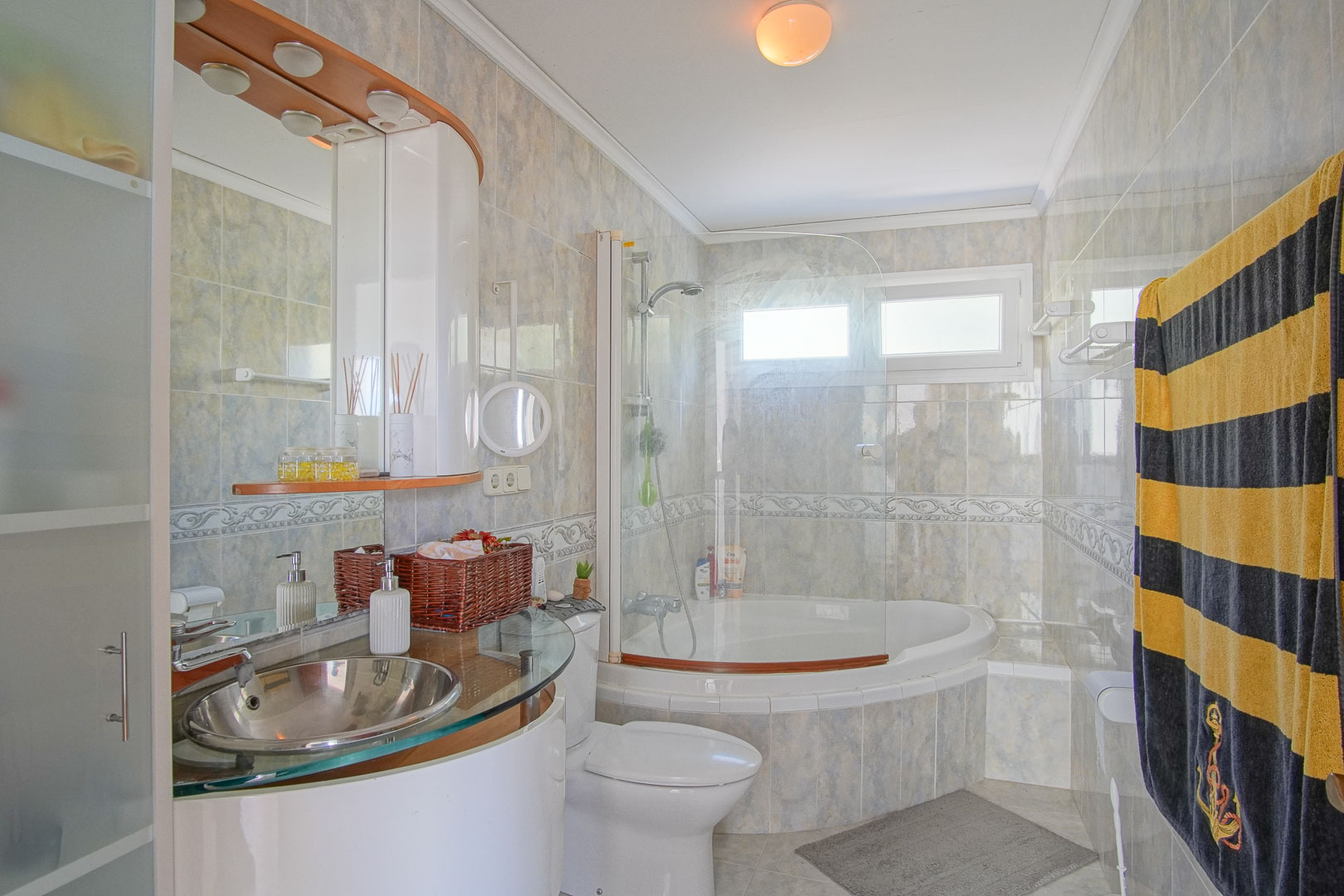 Komplettes Badezimmer mit Eckbadewanne, Glasduschwand und modernem Edelstahlwaschbecken auf einer Glasplatte. Geflieste Wände mit dekorativer Bordüre und Tageslicht.