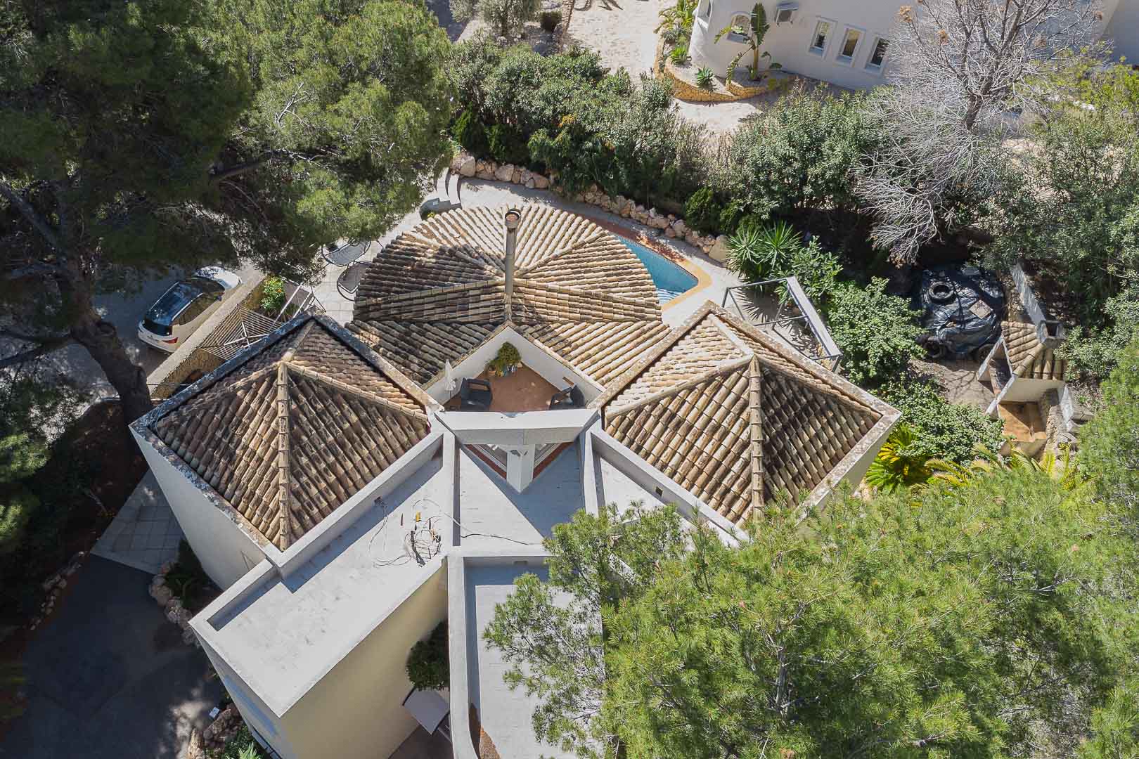Luftaufnahme der Villa mit markanten Pyramidendächern aus Ziegeln, einem zentralen Schornstein und einer blickgeschützten Dachterrasse inmitten mediterraner Pinien.