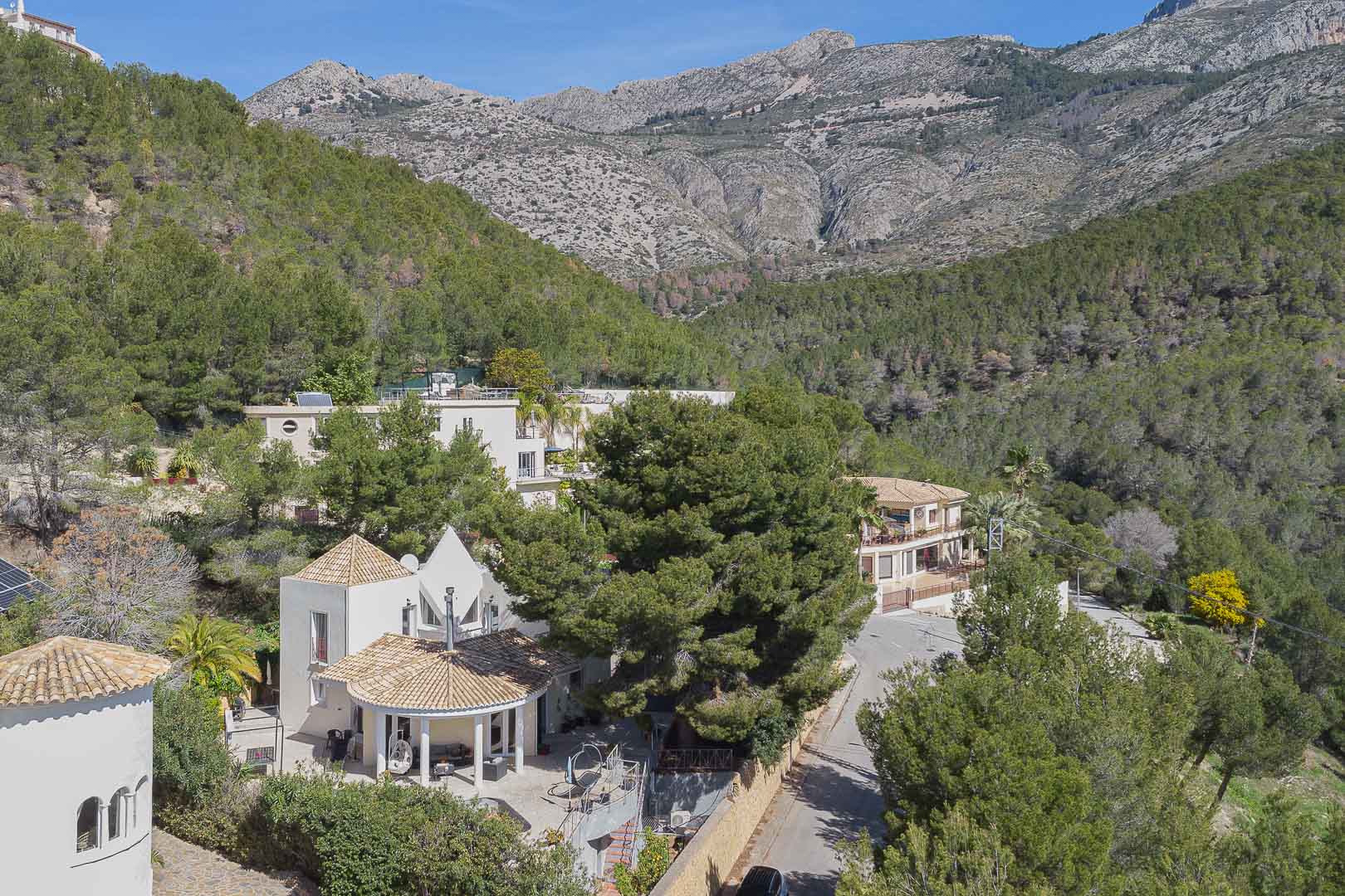 Luftaufnahme der Villa mit mediterraner Architektur und Ziegeldächern, eingebettet in einen Pinienwald vor der Kulisse der imposanten Berglandschaft.