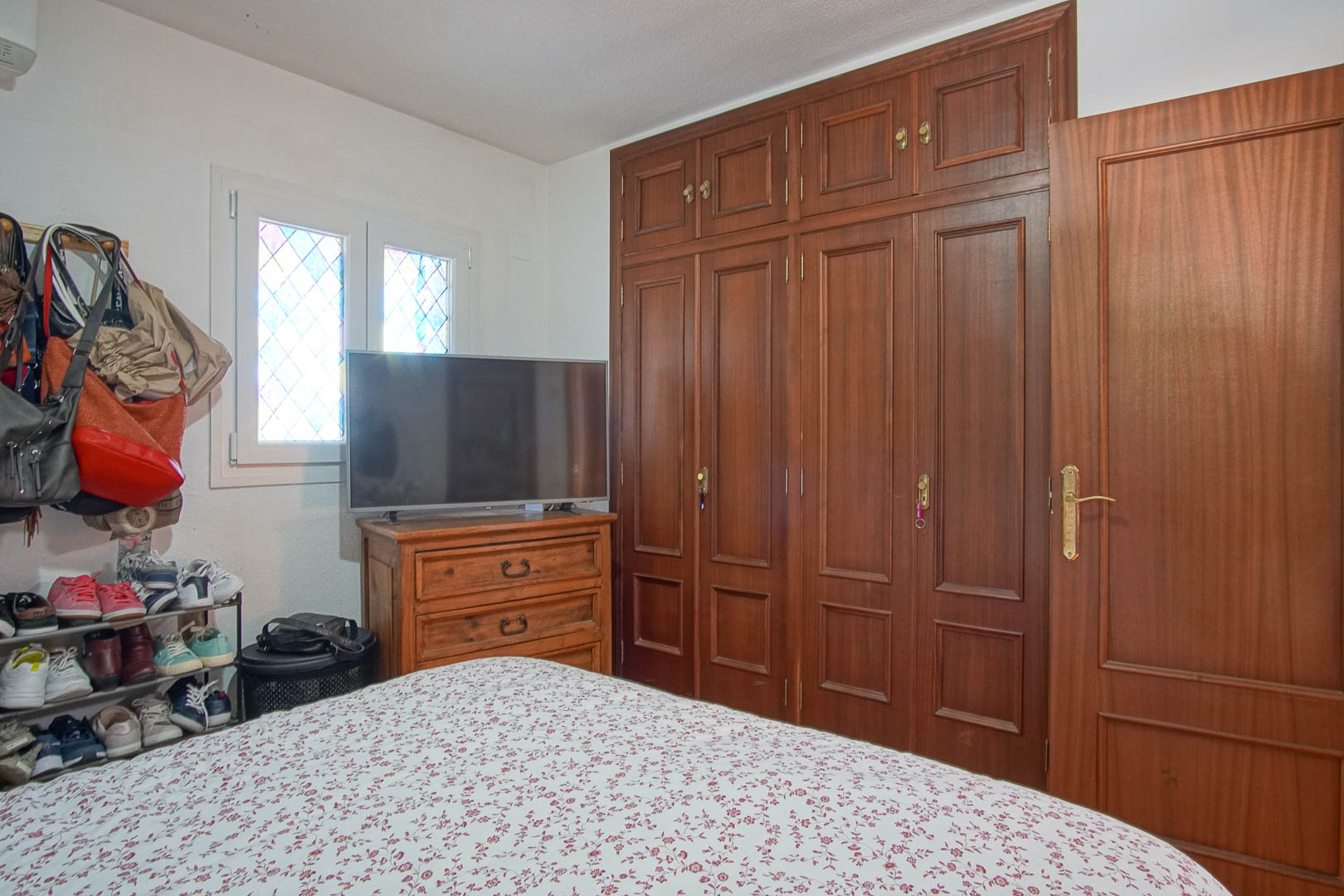 Schlafzimmer mit großem Einbauschrank aus dunklem Holz, passender Kommode mit Fernseher und einem Fenster mit Bleiglas-Elementen.