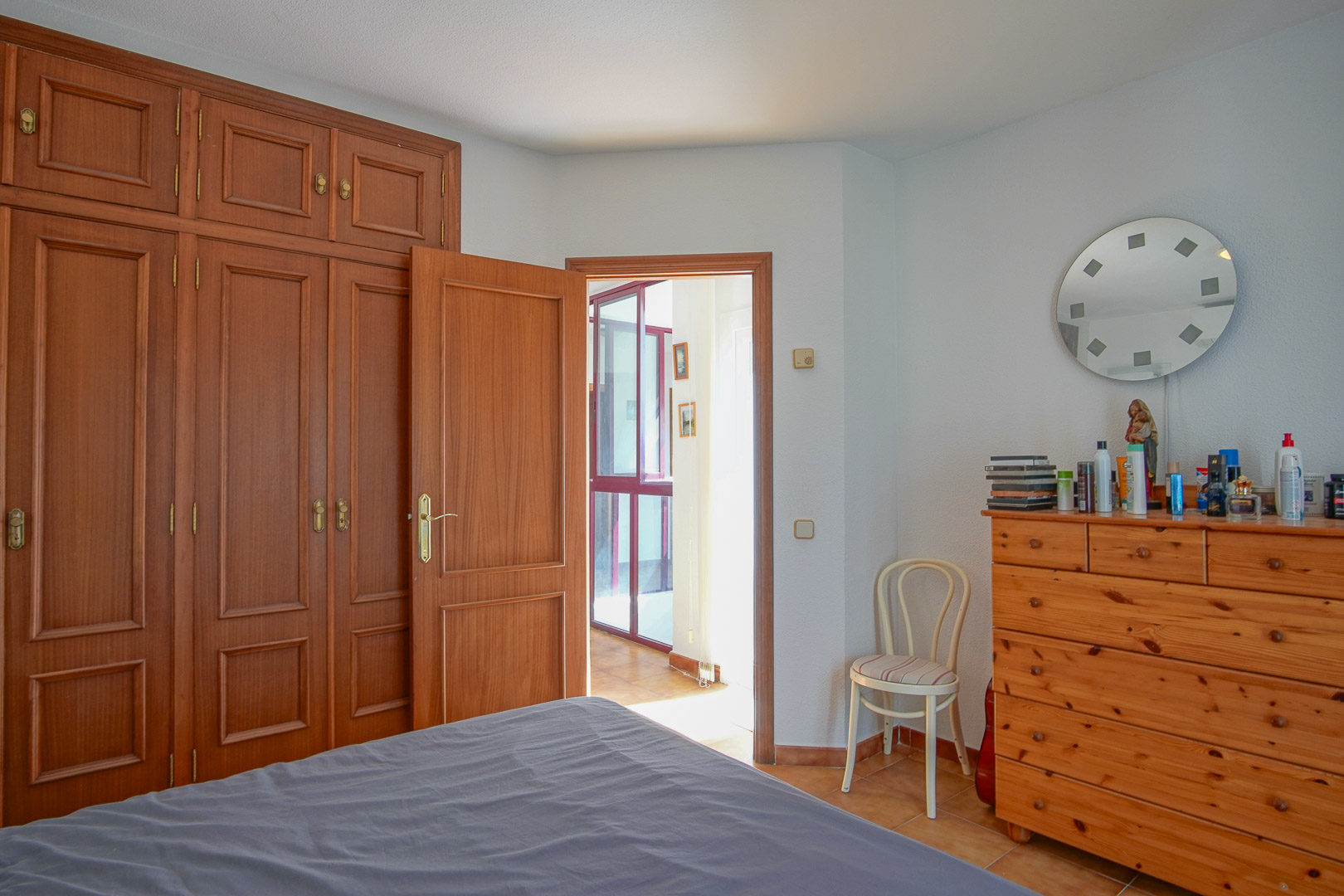 Schlafzimmer mit raumhohen Einbauschränken aus Holz, einer passenden Kommode und Fliesenboden. Der Raum bietet Zugang zu einem hellen Flur und besticht durch klassische Holzelemente.