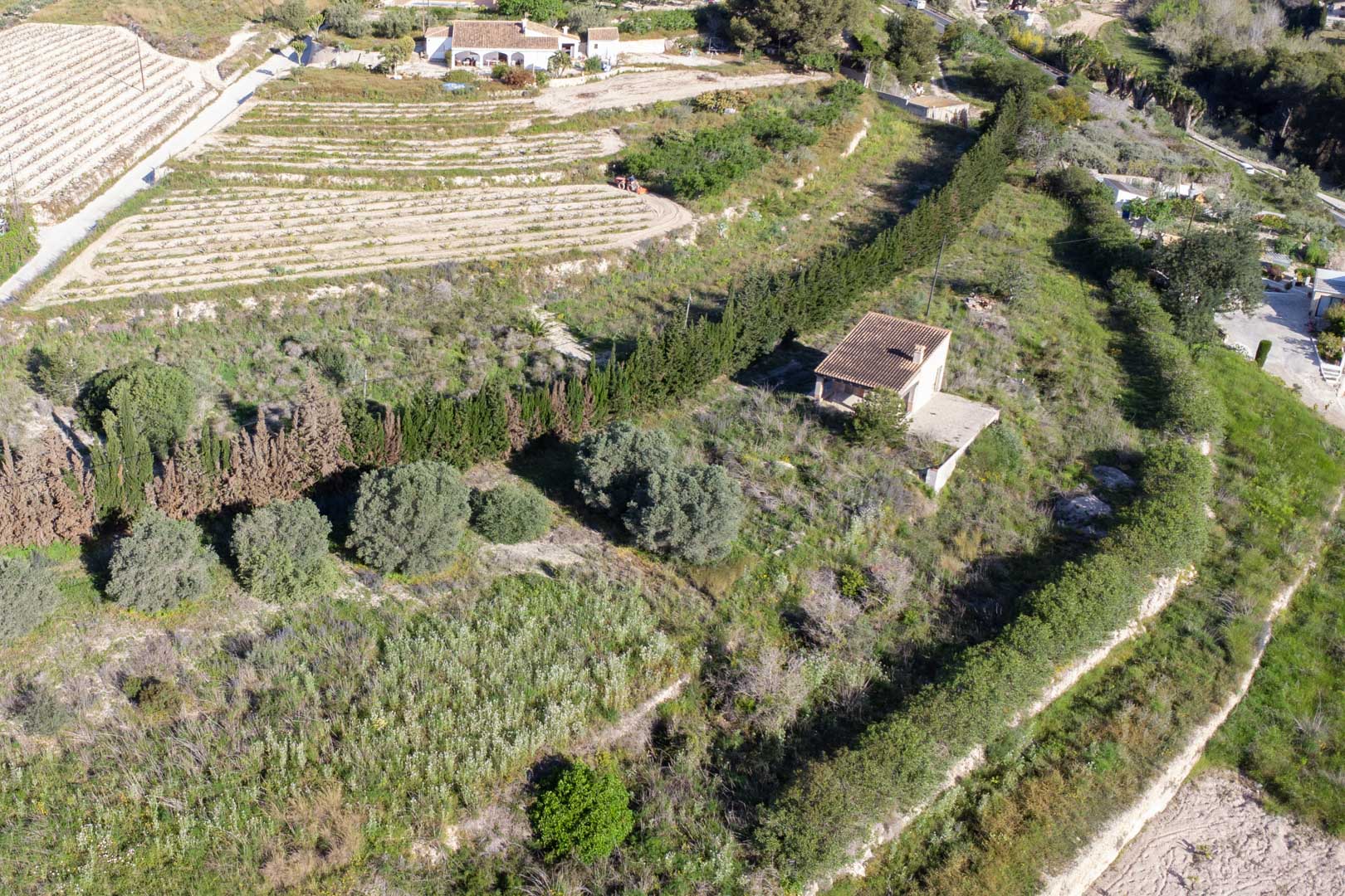 Luftaufnahme des 10.920 m2 großen Grundstücks in Teulada mit terrassiertem Gelände, mediterraner Vegetation und einem bestehenden ländlichen Nebengebäude.