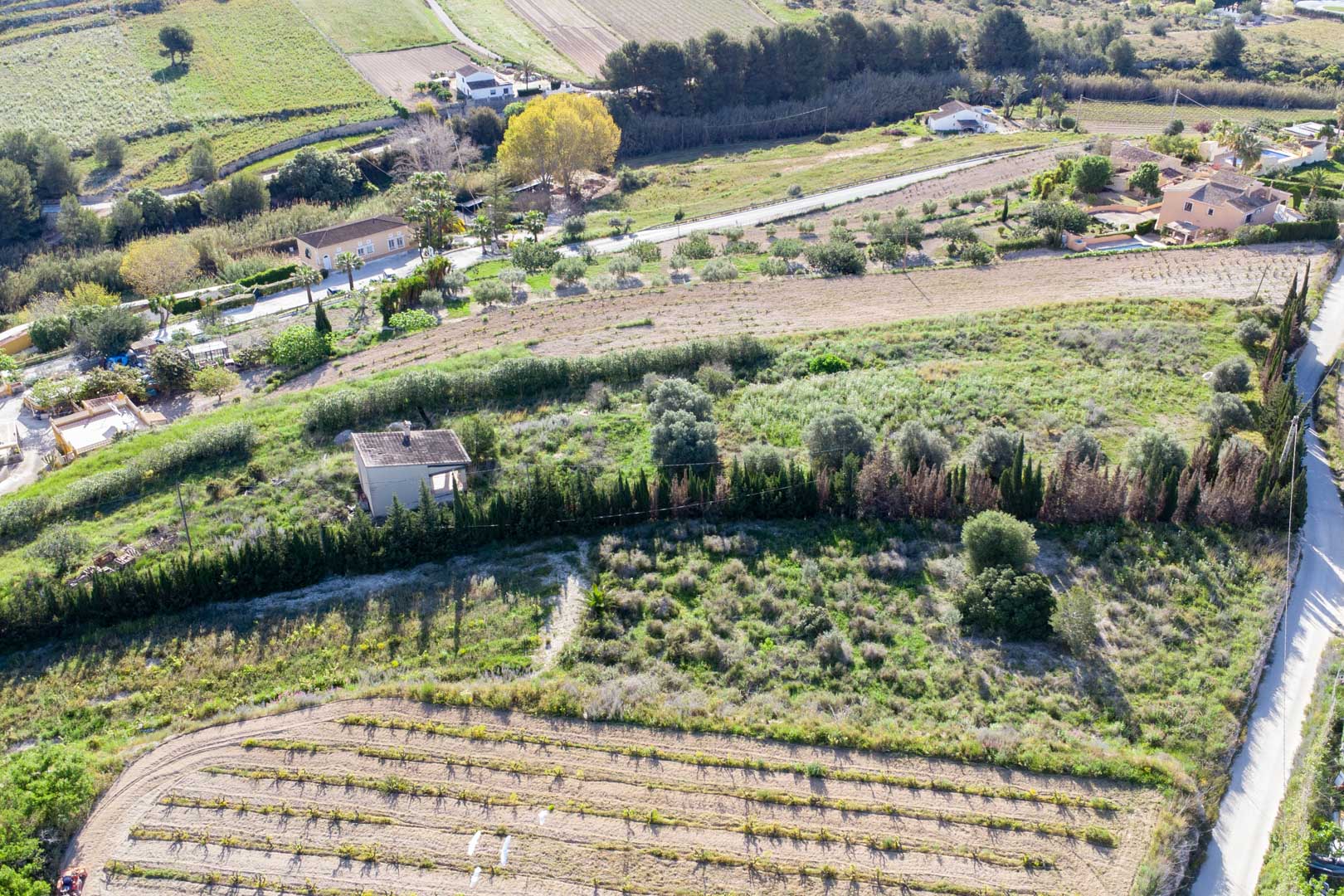 Luftaufnahme des weitläufigen rustikalen Grundstücks in Teulada mit terrassiertem Gelände, mediterraner Vegetation und einem kleinen Bestandsgebäude.