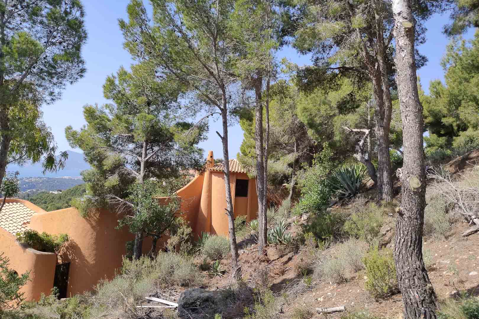 Außenansicht der organischen Architektur in Terrakottatönen, eingebettet in einen dichten Pinienwald und mediterrane Vegetation mit Meerblick.