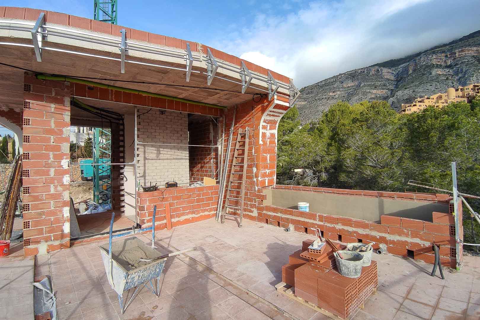 Bauphase der oberen Terrasse mit rotem Mauerwerk, Betonplatten und Metallhalterungen für die Fassade, umgeben von Pinien und Bergblick.