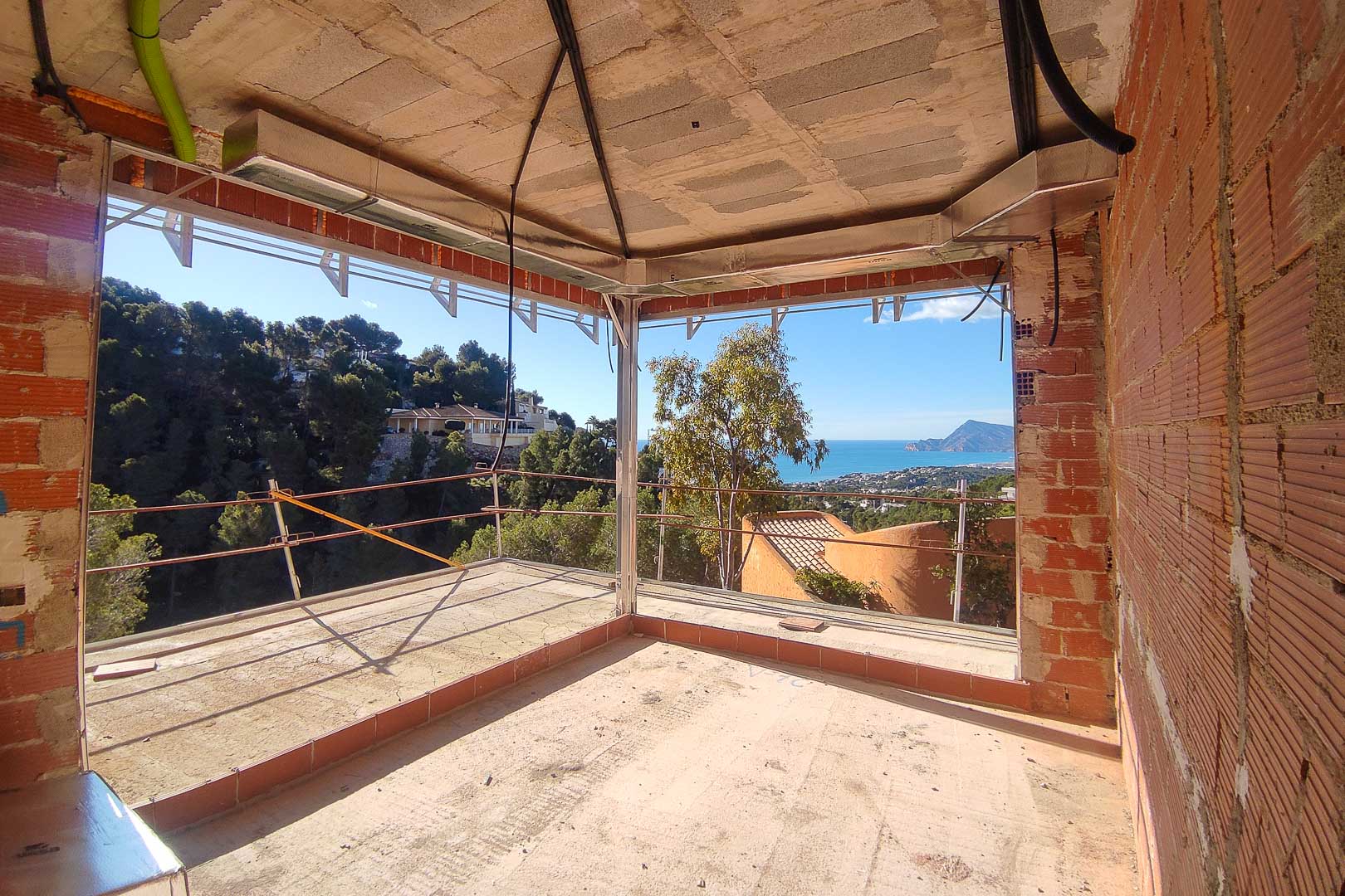 Panoramablick auf das Meer und die Berge aus einem Rohbau mit großen Eckfensterausschnitten und sichtbaren Klimatisierungsleitungen.