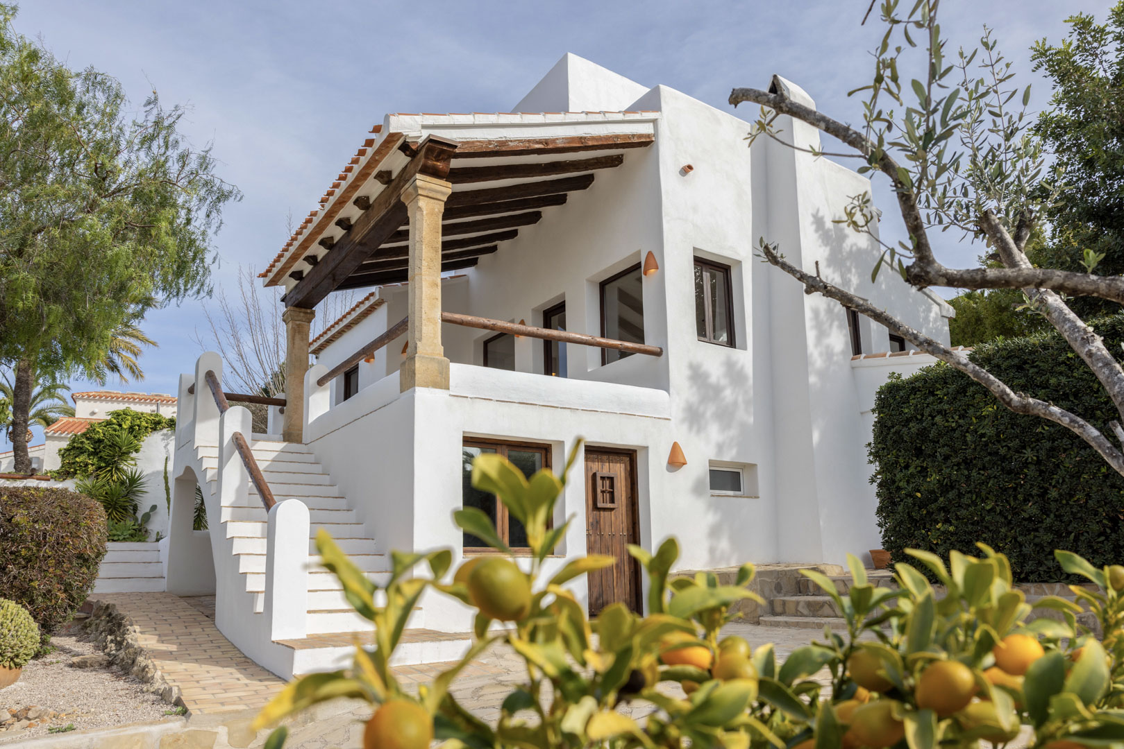 Außenansicht der Villa in Jávea, die die weiße Fassade mit überdachter Terrasse mit Holzbalken und Treppenzugang hervorhebt.