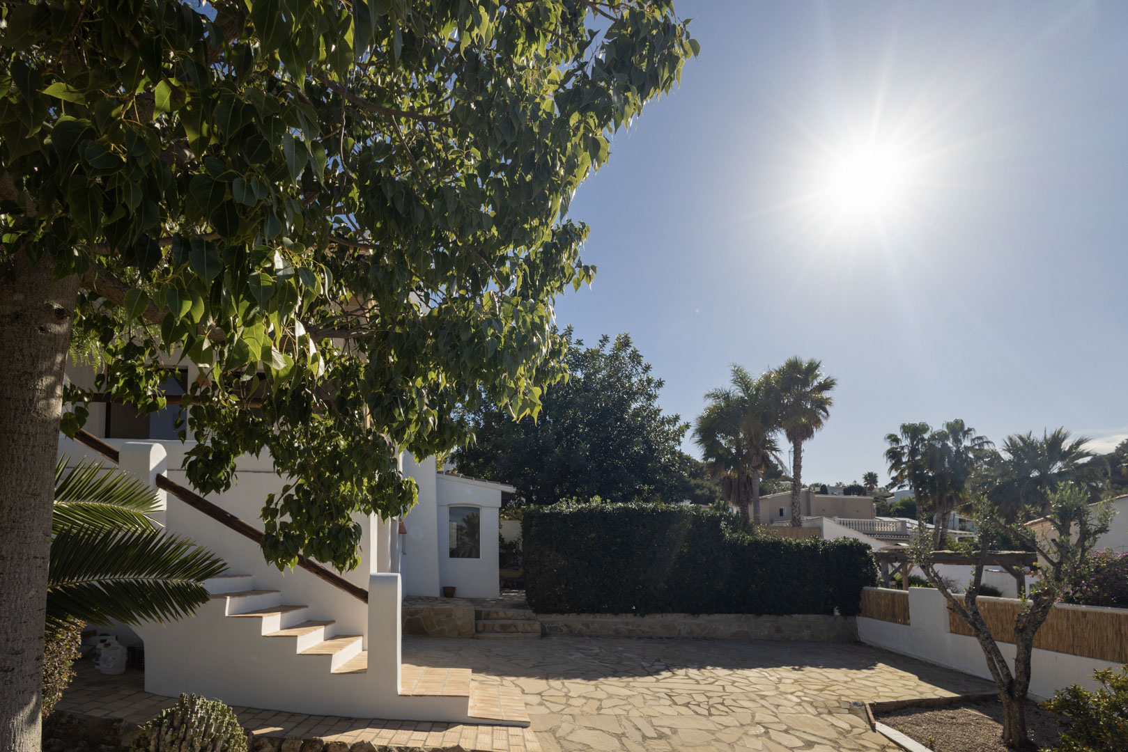 Außenansicht der Villa mit Steinterrasse, weißer Treppe und üppigem Grün unter der Sonne.