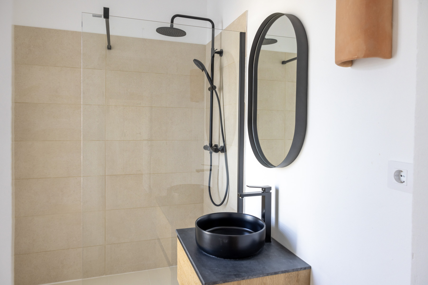 Badezimmerdetail mit begehbarer Dusche mit beigen Fliesen, schwarzem Aufsatzwaschbecken auf Schieferplatte und ovalem Spiegel.