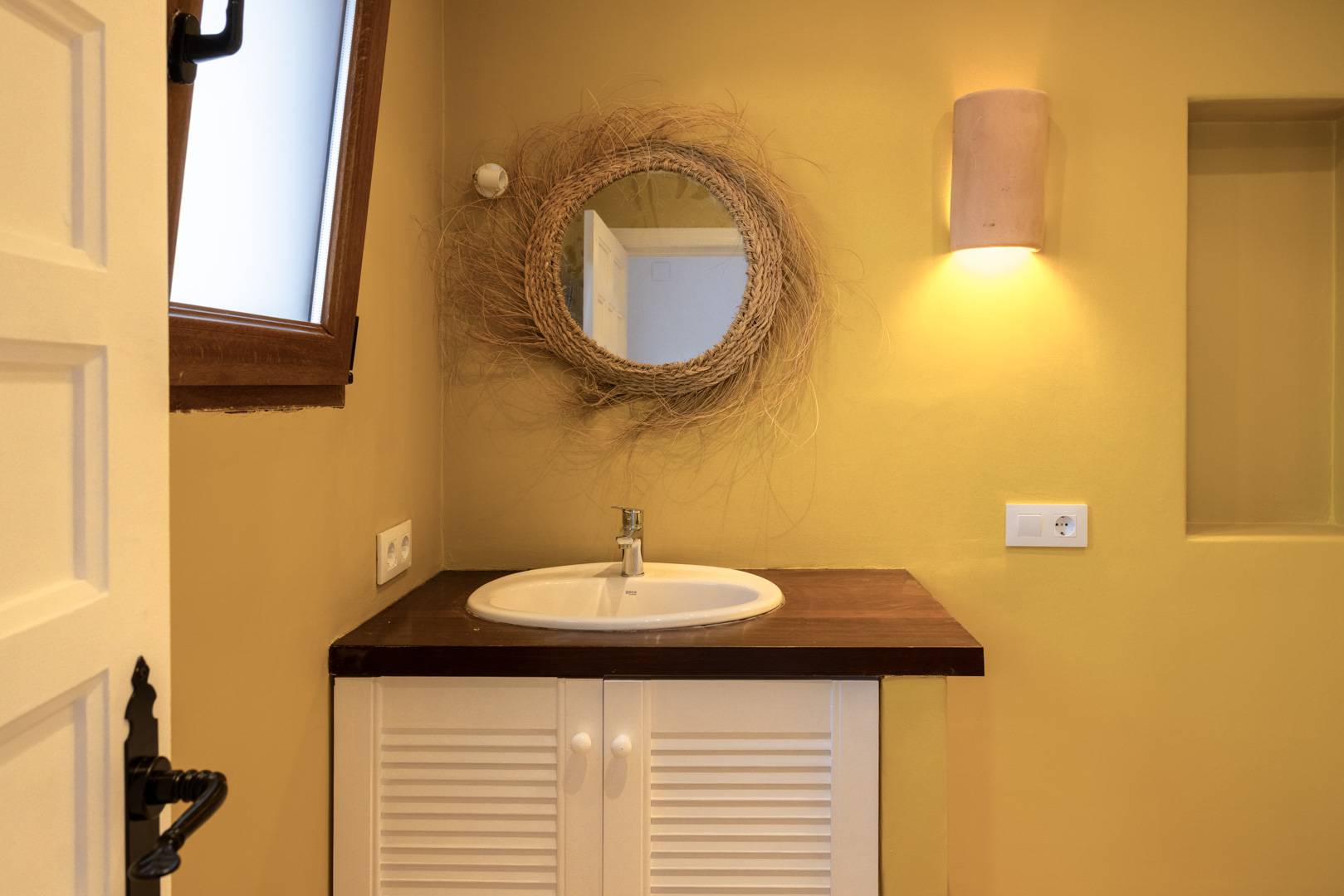 Badezimmerdetail mit ovalem Waschbecken, Weidenspiegel und dunkler Holzablage auf einem weißen Schrank.