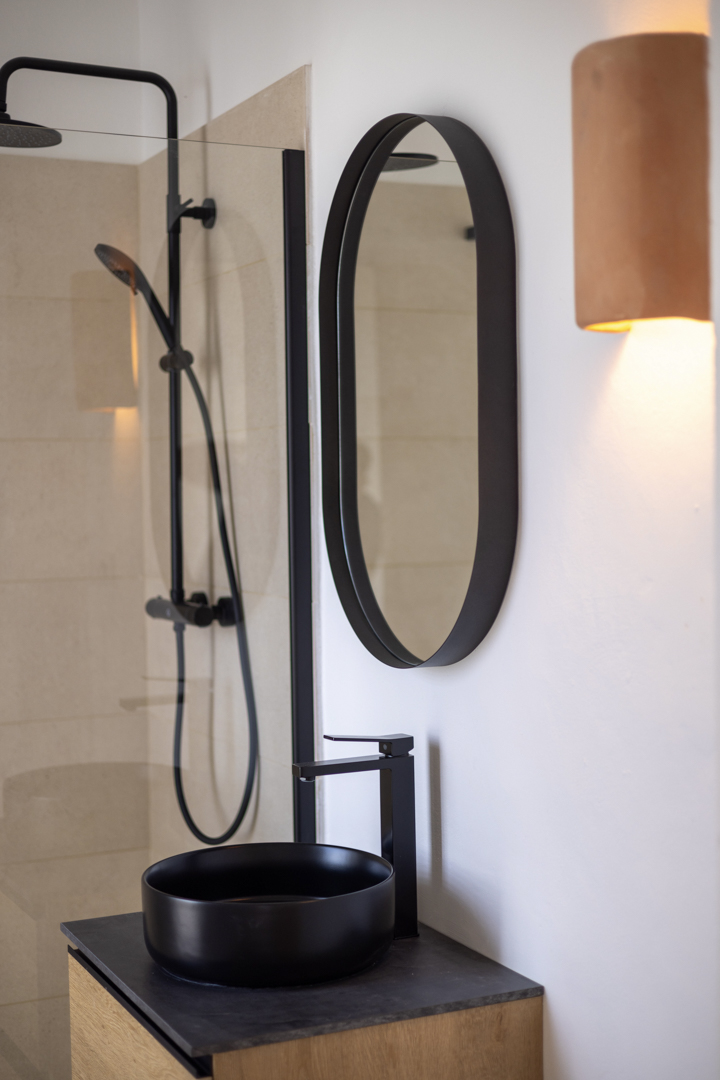 Badezimmerdetail mit rundem, schwarzem Waschbecken, ovalem Spiegel und Glasdusche.