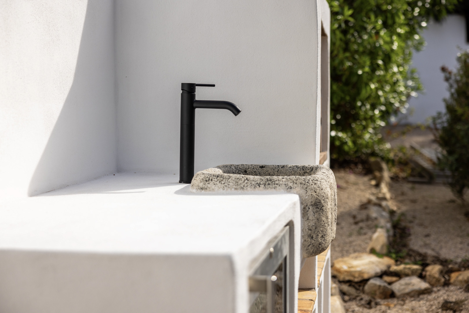 Detail der Außenküche: rustikales Steinwaschbecken mit schwarzem Wasserhahn auf einer weißen Arbeitsplatte.