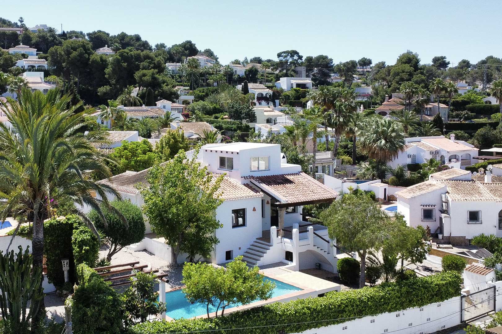 Erhöhte Ansicht einer weißen Villa mit türkisfarbenem Pool und Ziegeldach, eingebettet in üppiges Grün in Jávea.