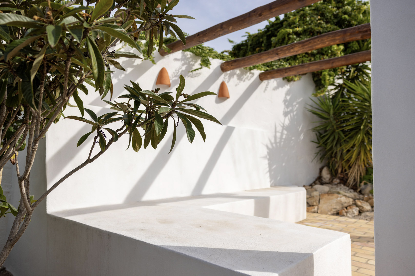 Terrassendetail mit weiß getünchter Betonbank, Holzpergola und üppigem Grün.