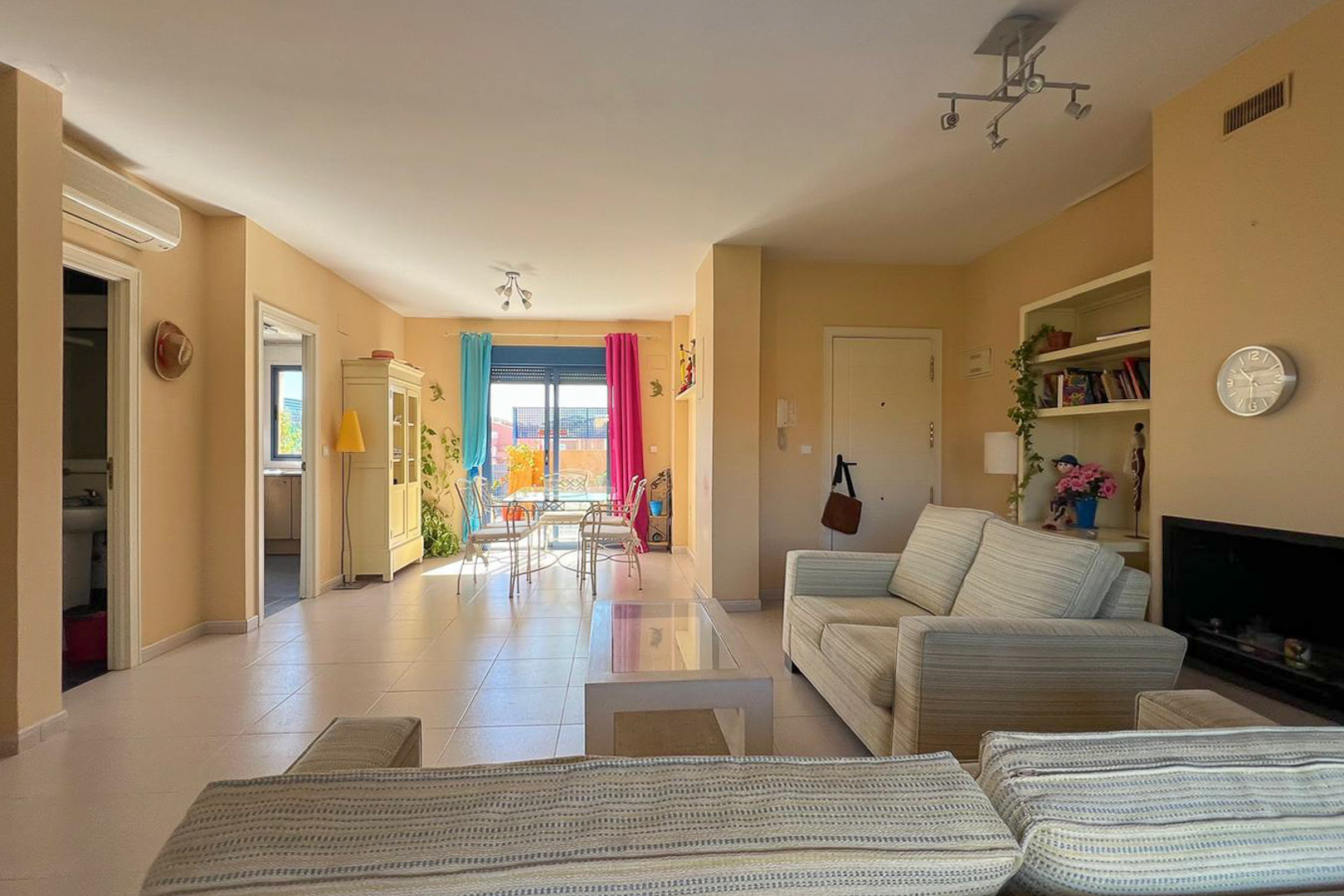 Helles Wohnzimmer mit Polstersofas und Zugang zu einer Terrasse mit farbenfrohen Vorhängen.