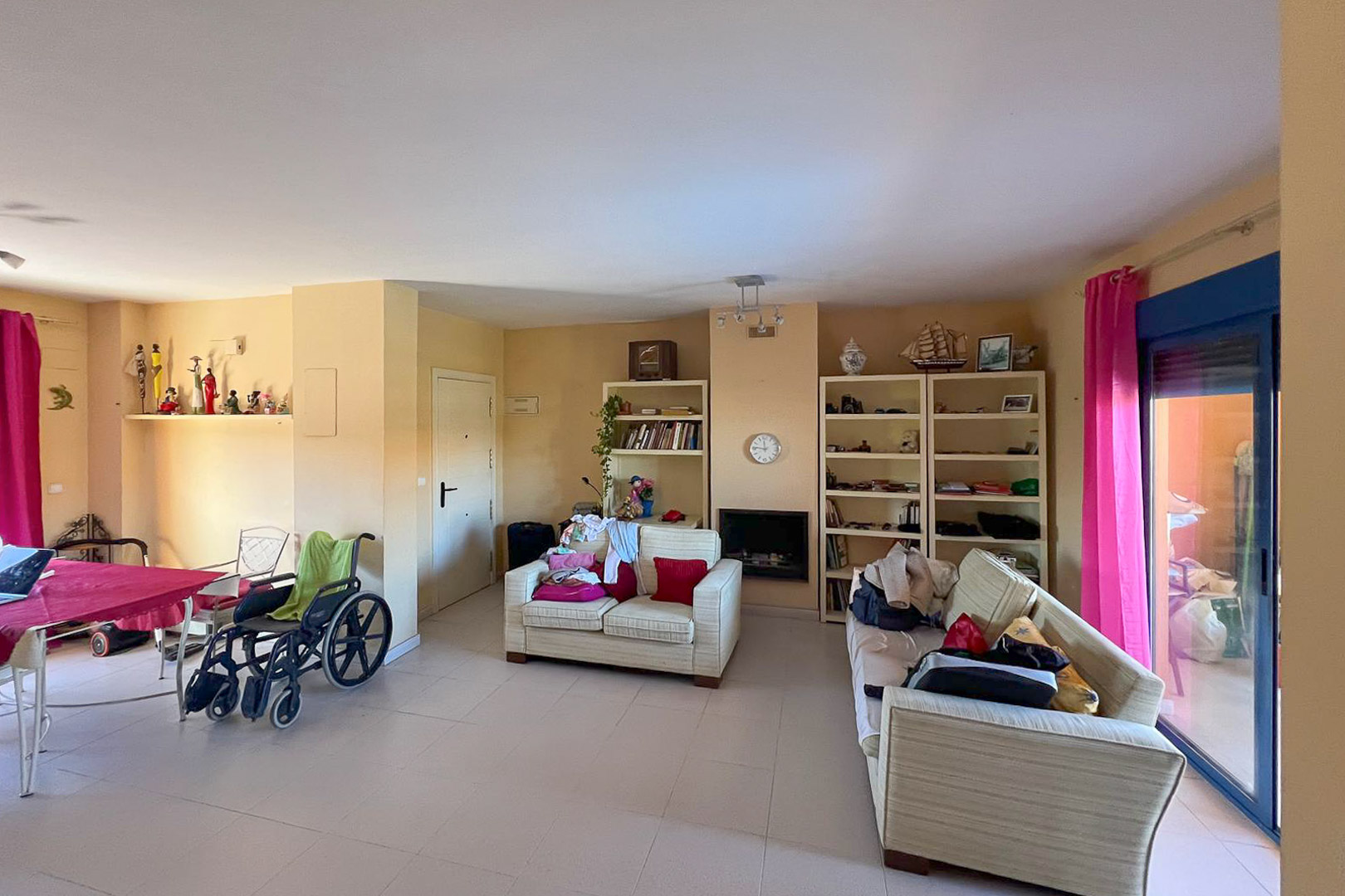 Wohn-Essbereich mit Regalen, Polstersofas und Terrassenzugang mit rosa Vorhängen.