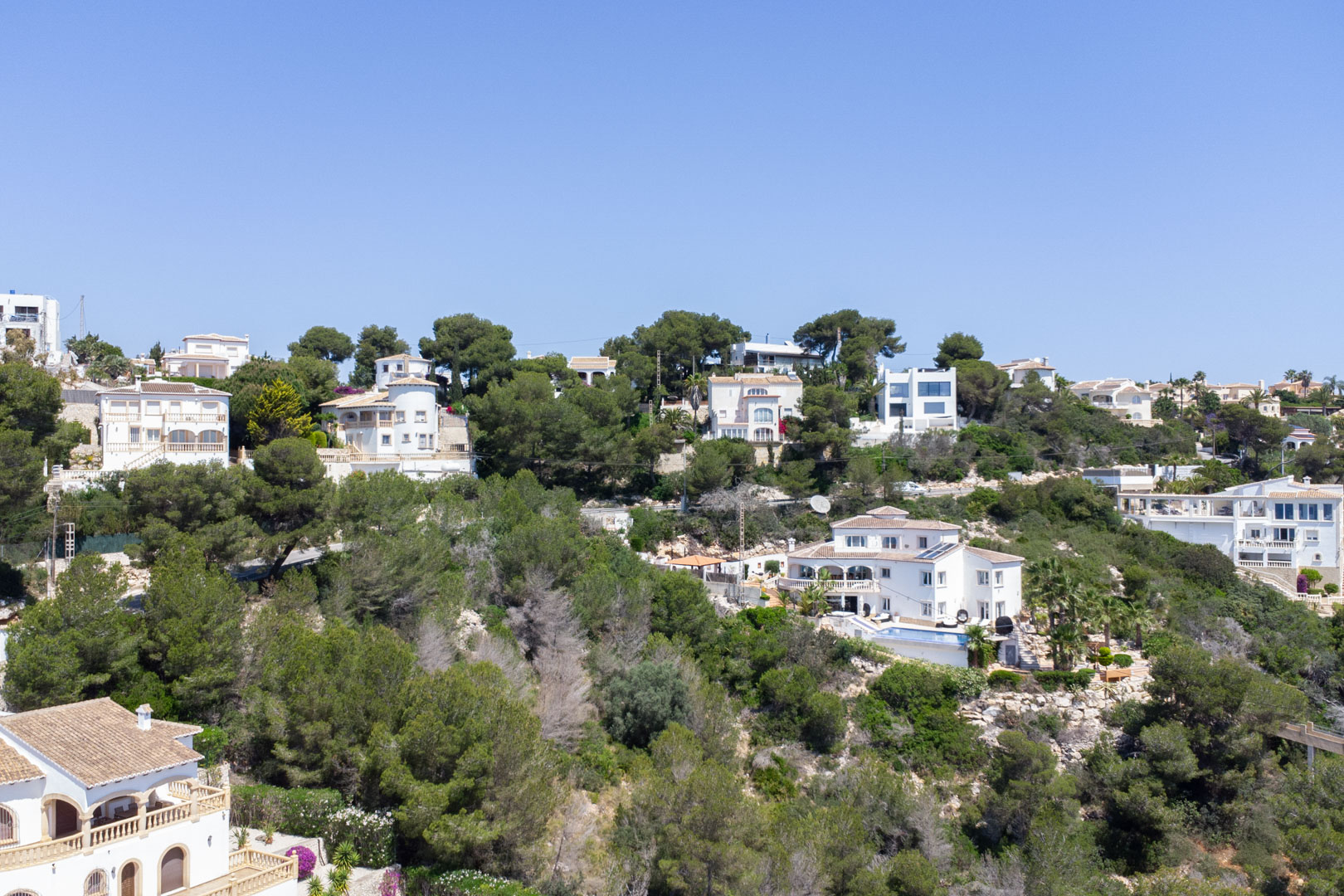 Erhöhte Ansicht des Wohngebiets Costa Nova in Jávea mit mediterranen Villen, die in einen bewaldeten Hang unter klarem blauem Himmel eingebettet sind.