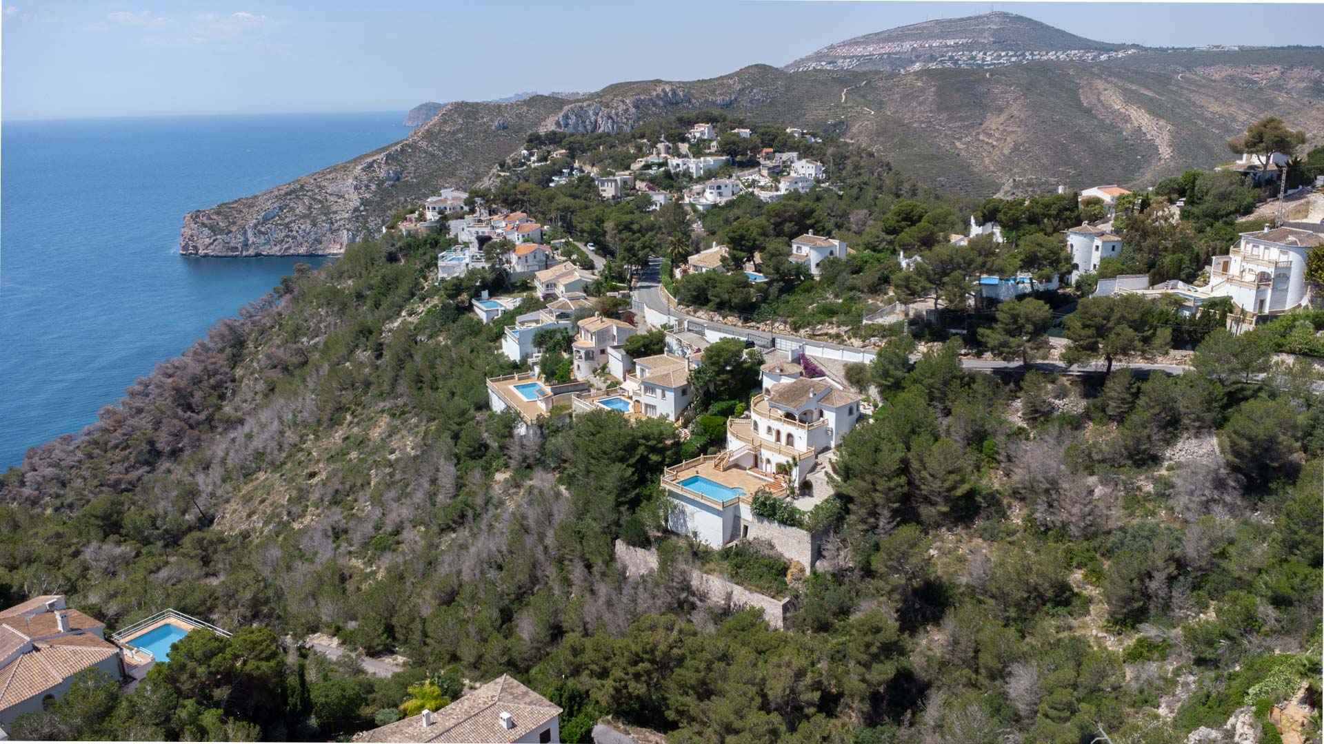 Luftaufnahme der Küste von Jávea mit Wohnvillen an einem bewaldeten Hang mit Blick auf das Mittelmeer und die zerklüfteten Klippen.