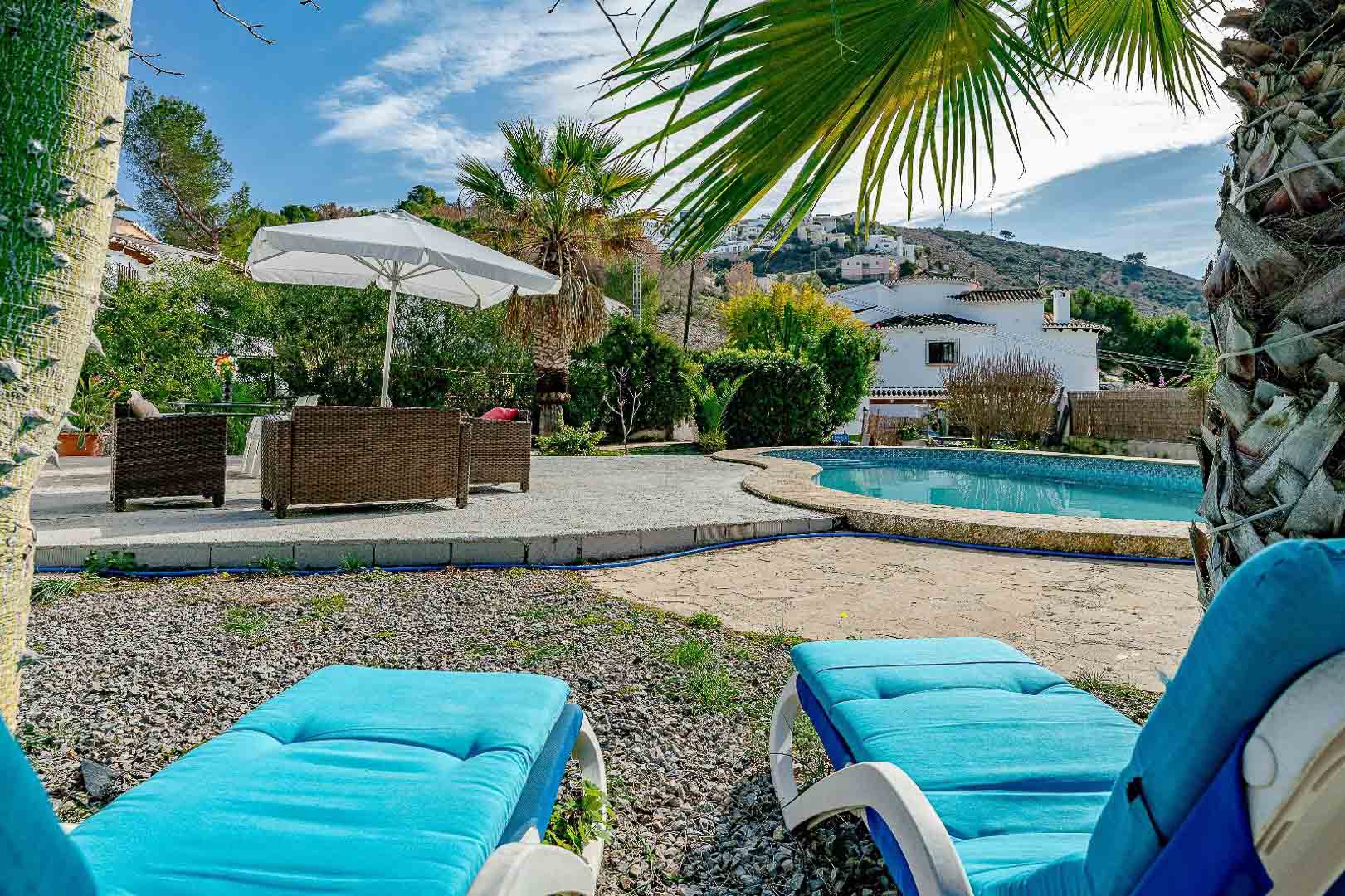 Außenpoolbereich mit nierenförmigem Becken, Steinterrasse, Korbmöbeln und blauen Sonnenliegen, eingebettet in eine mediterrane Gartenlandschaft mit Palmen.