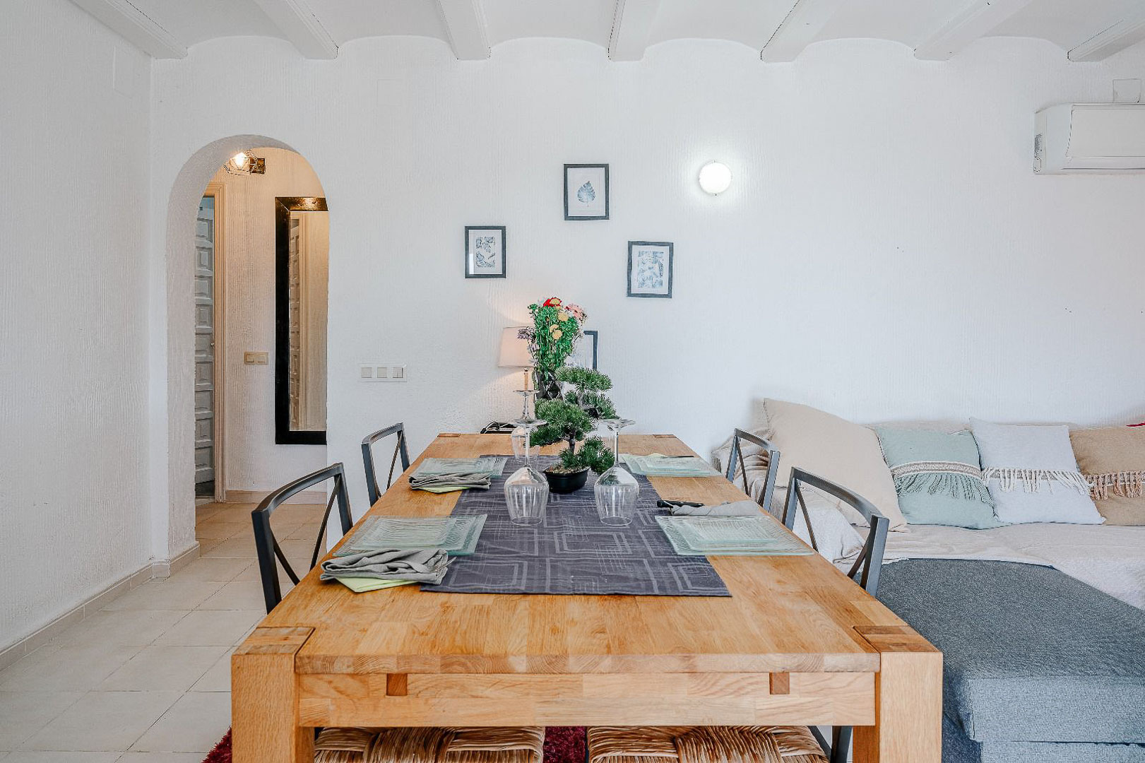 Heller Essbereich mit Massivholztisch und weißer Balkendecke. Ein Rundbogen führt zum Flur, angrenzend an den Wohnbereich mit Sofa und Klimaanlage.