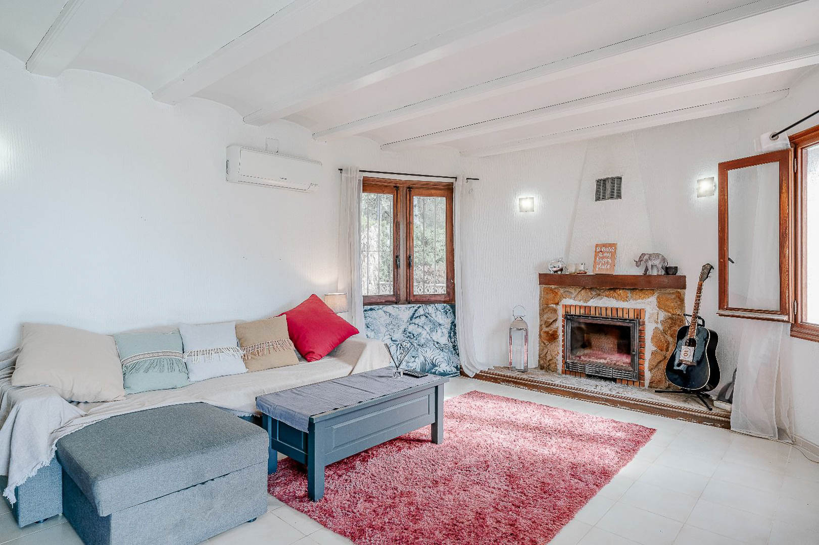 Heller Wohnbereich mit weißen Holzbalkendecken, einem rustikalen Natursteinkamin und Klimaanlage. Holzfenster sorgen für reichlich natürliches Licht im mediterranen Raum.