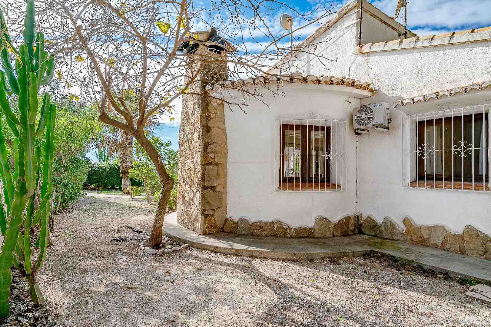 Mediterrane Fassade mit Natursteinkamin, Holzfenstern mit weißen Sicherheitsgittern und einem pflegeleichten Garten mit Kies und einheimischen Pflanzen.