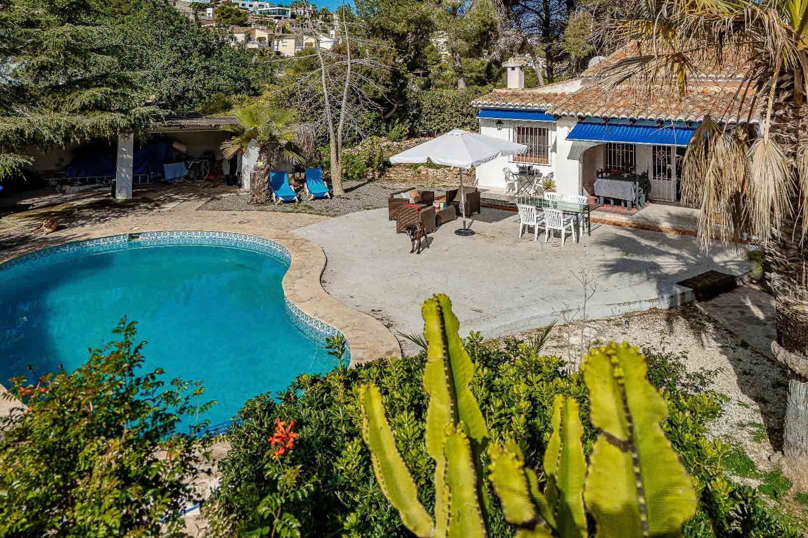 Mediterraner Außenbereich mit nierenförmigem Pool, großzügiger gepflasterter Terrasse mit Essbereich und weißer Fassade mit blauen Markisen und Ziegeldach.