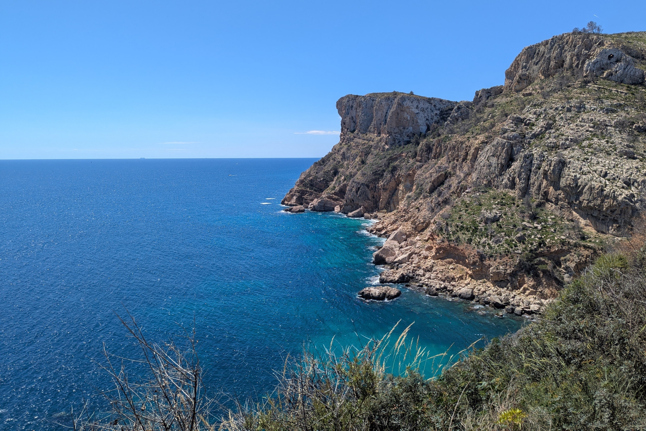 Panoramablick auf die zerklüfteten Kalksteinfelsen und das türkisfarbene Mittelmeerwasser bei Cala Llebeig, umgeben von typischer Küstenvegetation.
