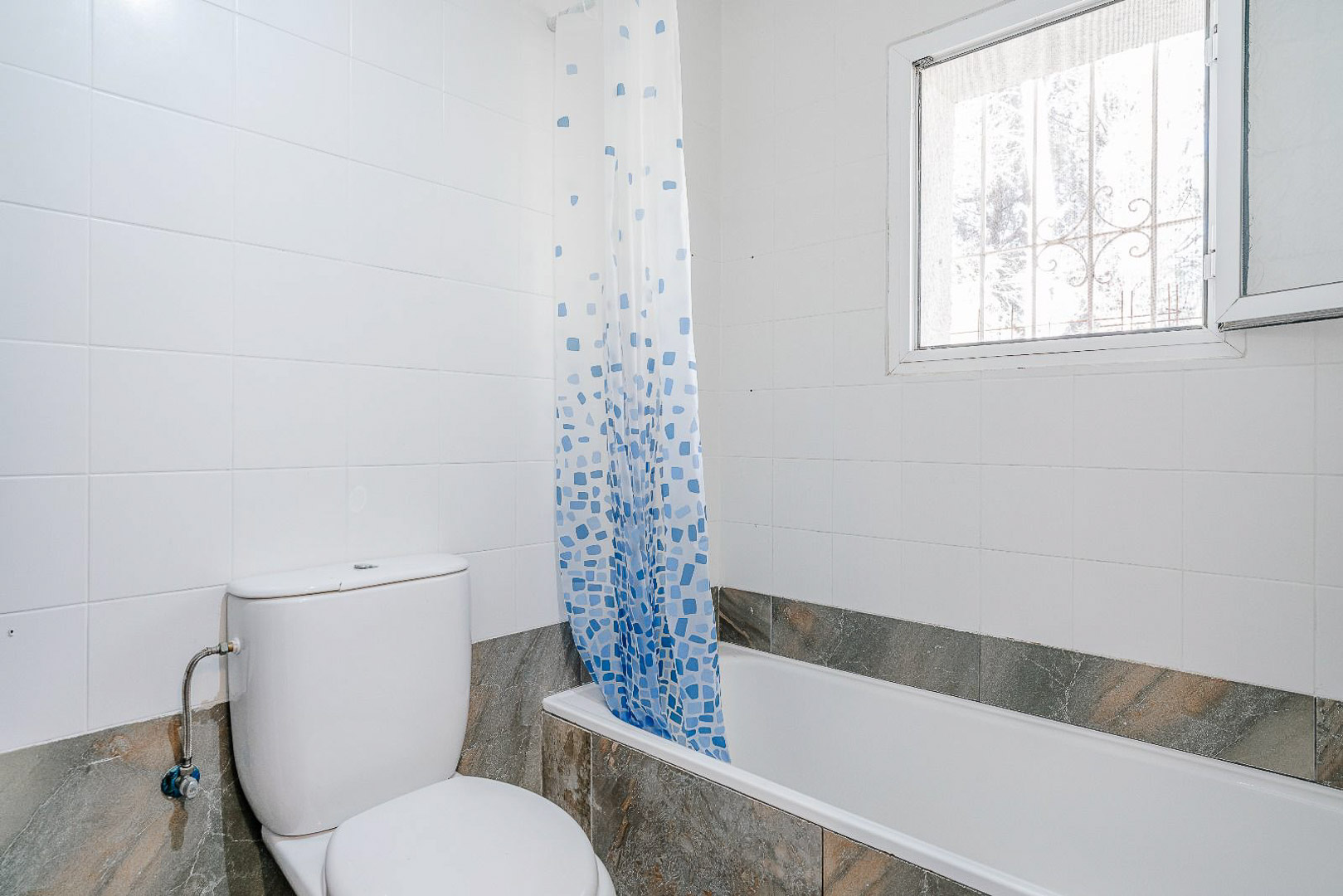 Renoviertes Badezimmer mit Einbaubadewanne, weißem WC und hellen Wandfliesen. Ein Fenster mit Sicherheitsgitter sorgt für natürliches Licht und Belüftung.