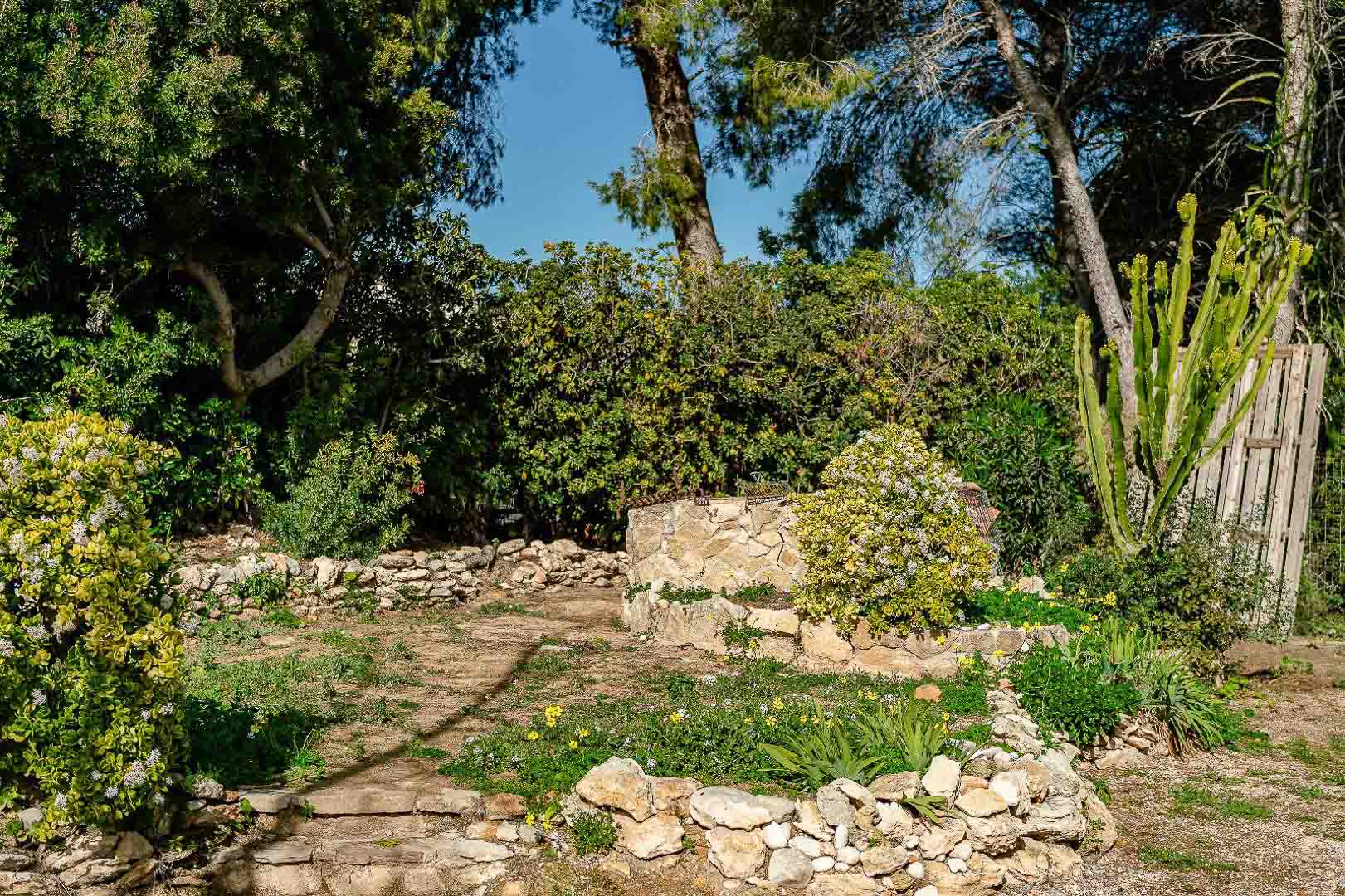 Rustikaler Garten mit Trockensteinmauern, mediterraner Bepflanzung und einem zentralen Steinbrunnen inmitten von Pinien und Sträuchern.