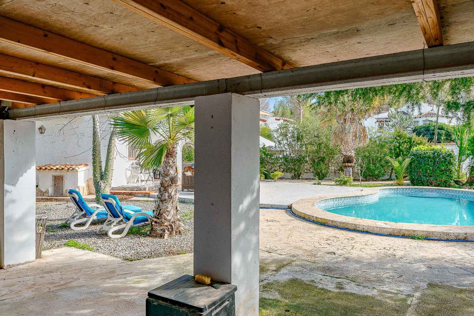 Überdachte Veranda mit Holzbalkendecke und Blick auf den nierenförmigen Pool sowie den mediterranen Garten mit Palmen und Sonnenliegen.