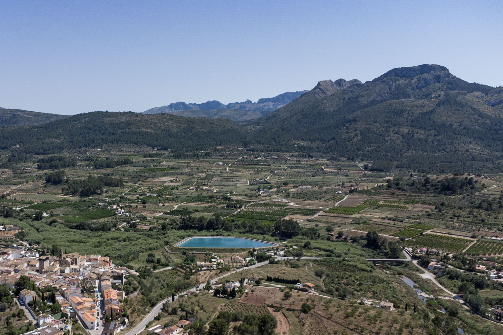 Panorama-Luftaufnahme des Jalón-Tals mit dem Dorf Alcalalí, landwirtschaftlichen Flächen, einem Wasserreservoir und den umliegenden Gebirgsketten.