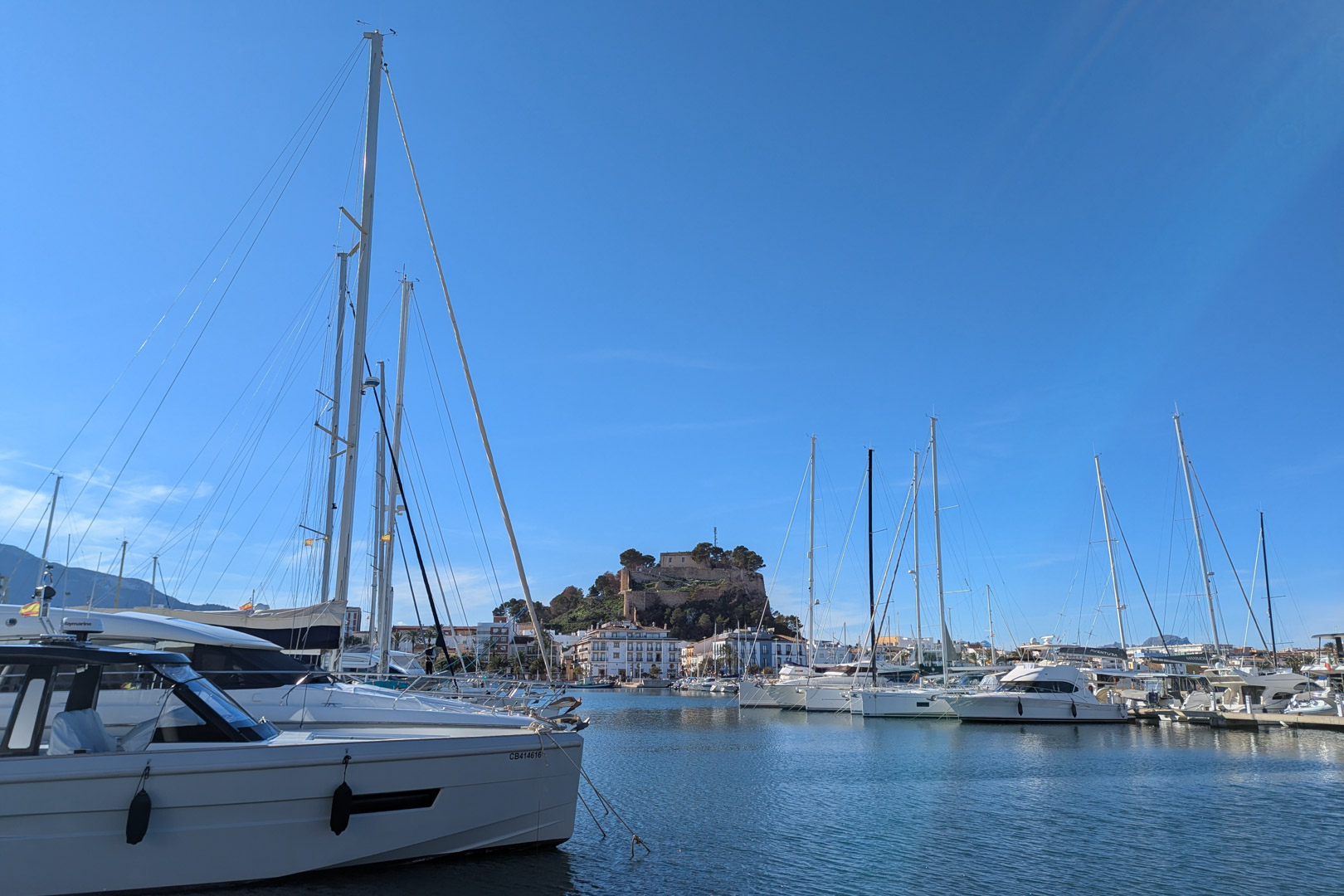 Blick auf den Yachthafen mit Yachten und der Burg von Dénia im Hintergrund unter einem klaren blauen Himmel.