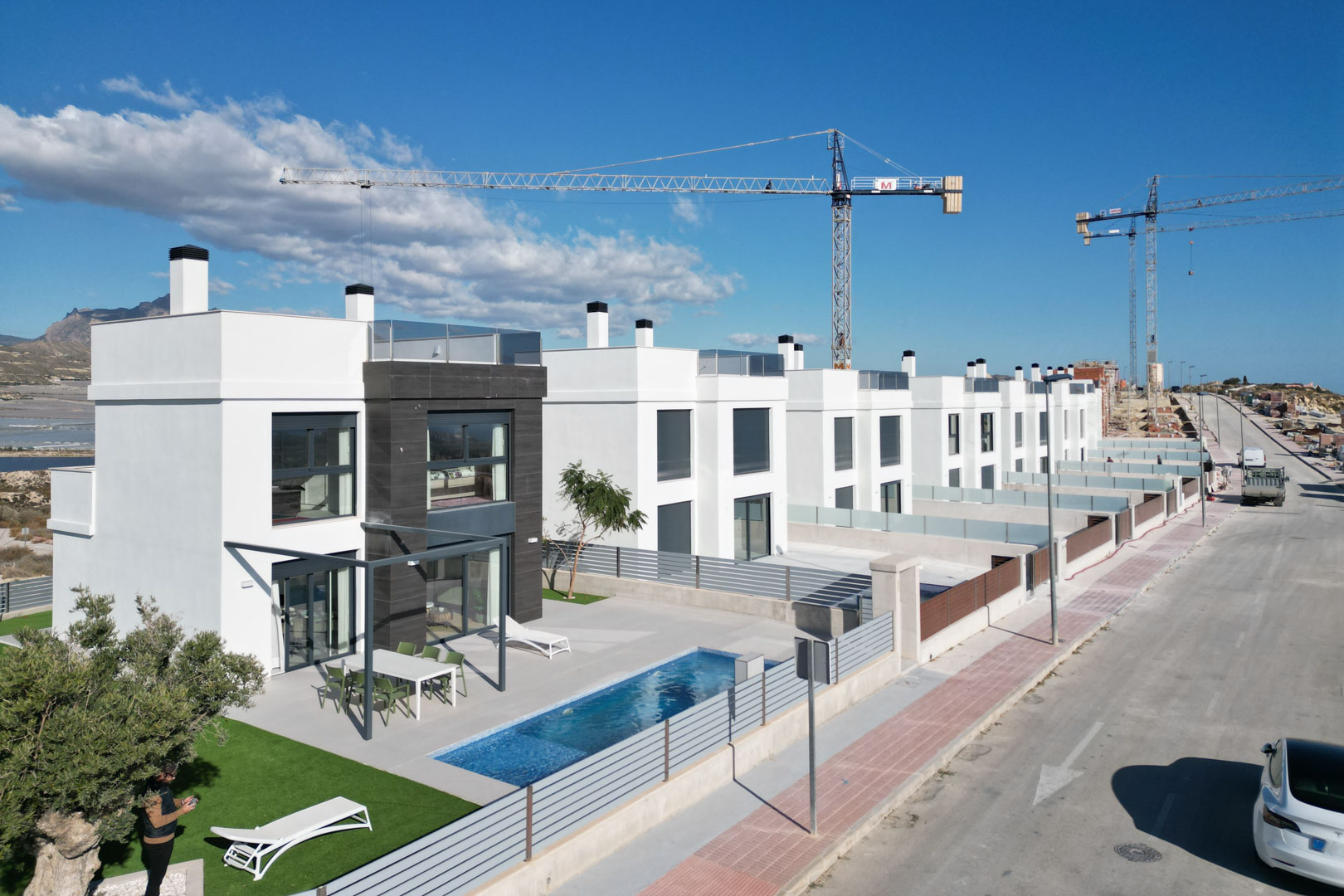 Luftaufnahme moderner Villen mit weißen Fassaden, dunklen Steinakzenten, privaten Pools, möblierten Terrassen und Dachterrassen in Hanglage.