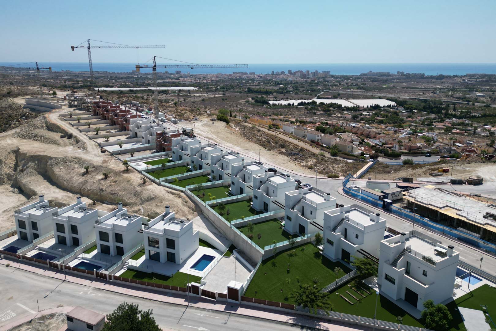 Luftaufnahme moderner weißer Villen in Hanglage mit privaten Gartenparzellen, individuellen Pools und weitem Blick über die mediterrane Küstenlandschaft.