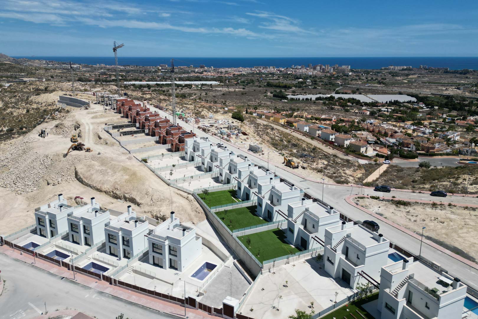 Luftaufnahme moderner weißer Villen in Hanglage mit privaten Pools, Gärten und Panoramablick auf das Mittelmeer und die Küstenlandschaft.