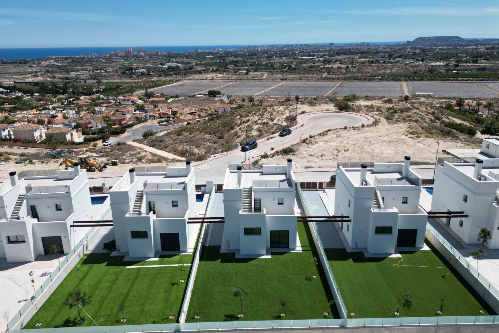 Luftaufnahme moderner weißer Villen mit privaten Rasenflächen und Dachterrassen, gelegen in erhöhter Position mit weitem Blick auf das Meer und die Küstenstadt.