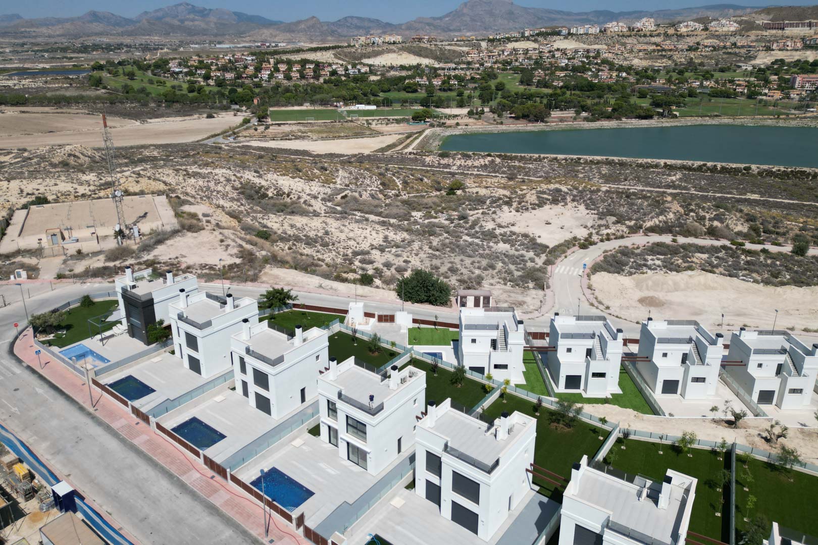 Luftaufnahme zeitgenössischer weißer Villen mit privaten Pools und Gärten, gelegen nahe einem Golfplatz und Stausee mit Bergblick.