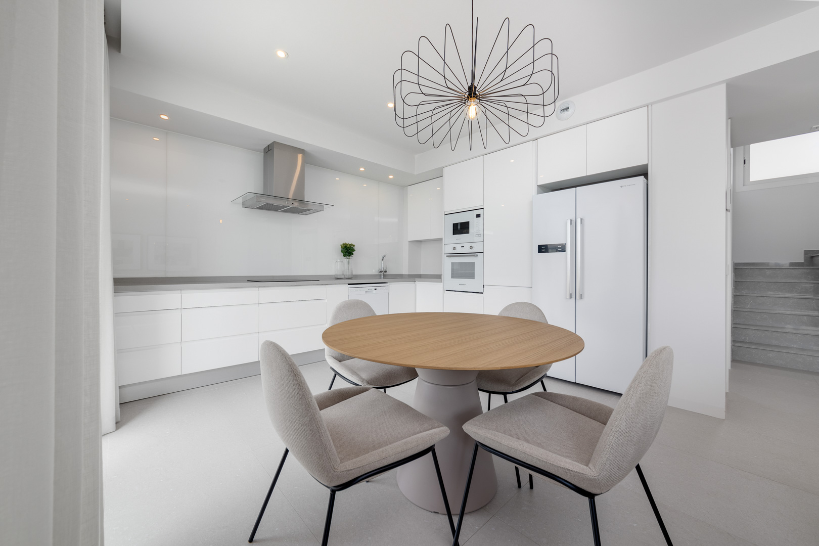 Minimalistische weiße Küche mit integrierten Geräten, grauer Arbeitsplatte und Essbereich mit rundem Holztisch und dekorativer Pendelleuchte.