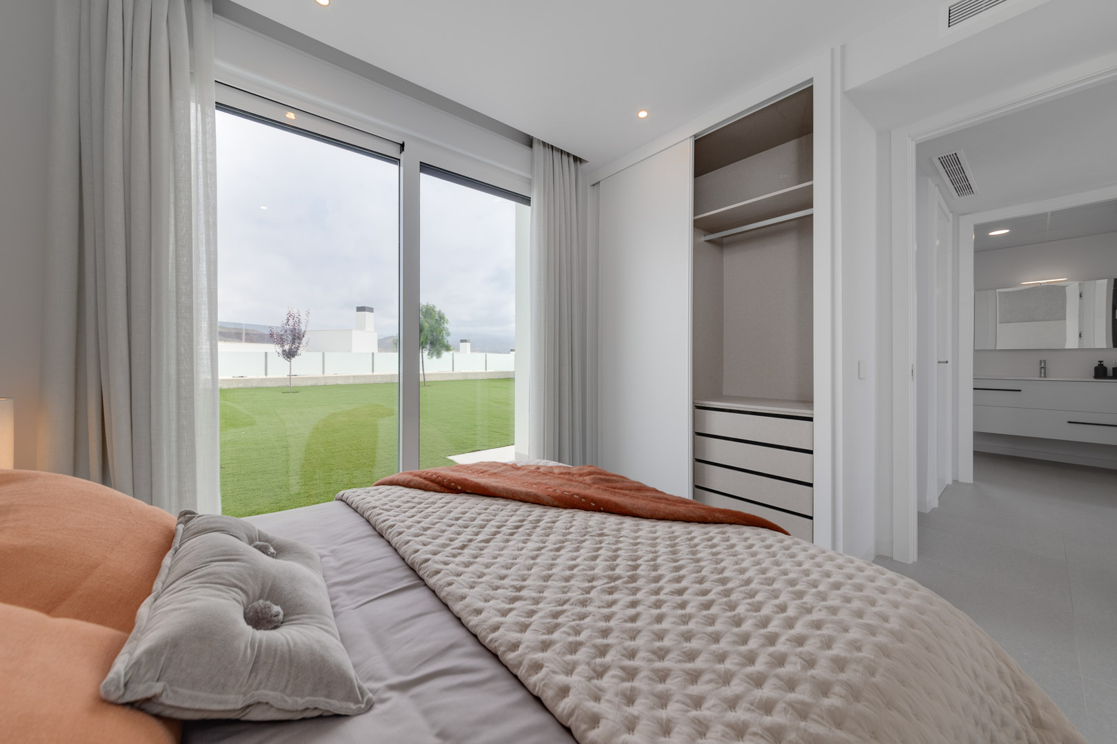 Schlafzimmer mit Einbauschrank, bodentiefen Fenstern mit direktem Gartenzugang und Blick in das en-suite Badezimmer mit hängendem Waschtisch.