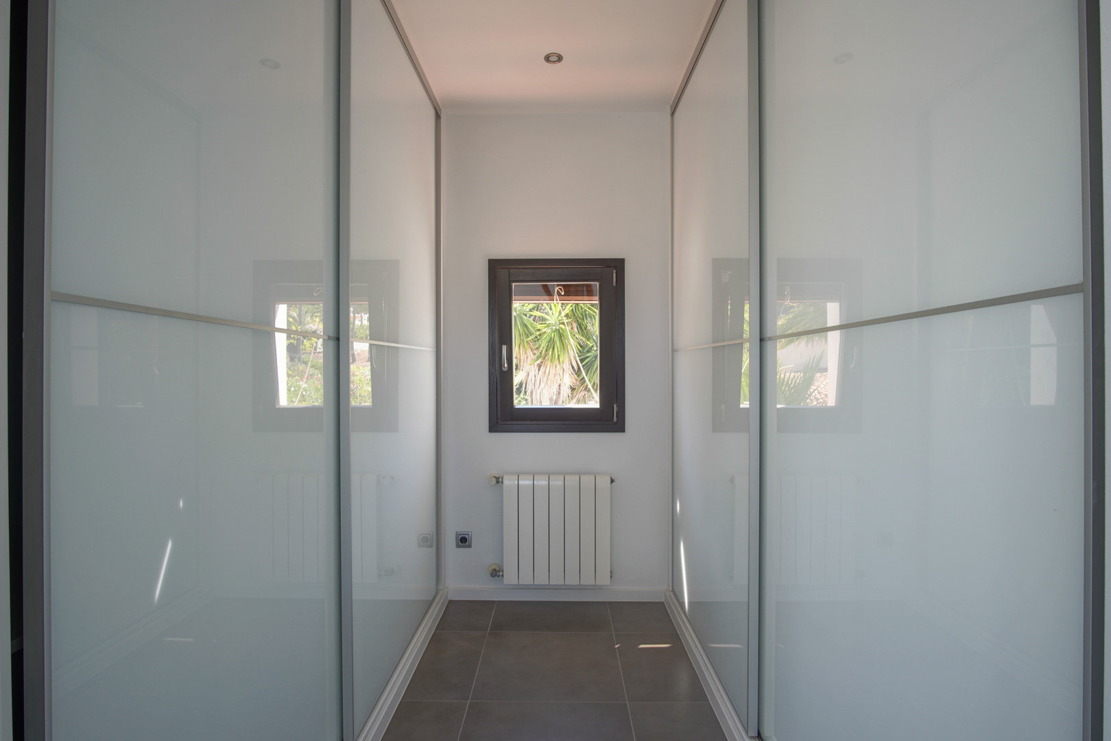 Ankleidezimmer mit Einbauschränken mit Schiebetüren aus Milchglas, einem Heizkörper und Fenster mit Blick auf die mediterrane Bepflanzung.