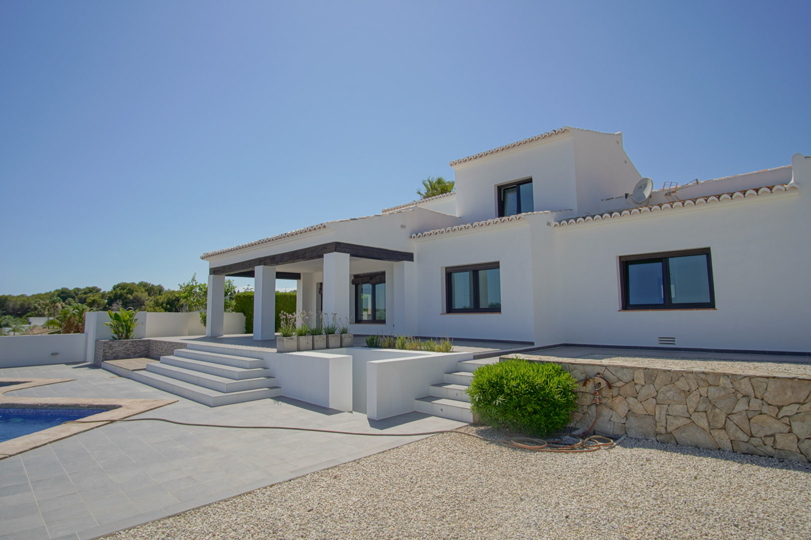 Außenansicht der mediterranen Villa mit weißer Fassade, dunklem Balken-Vordach und abgestufter Terrasse, die an den Poolbereich und Steinmauern grenzt.