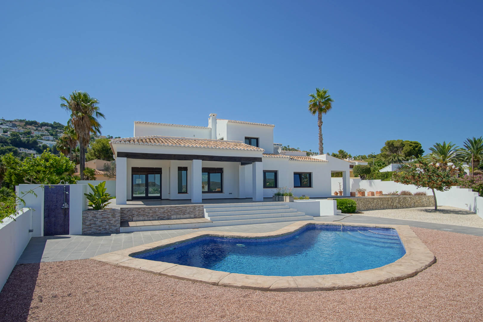 Außenansicht der Villa mit einem nierenförmigen Pool, pflegeleichtem Kiesgarten und einer überdachten Terrasse mit dunklen Holzbalken und weißen Säulen.