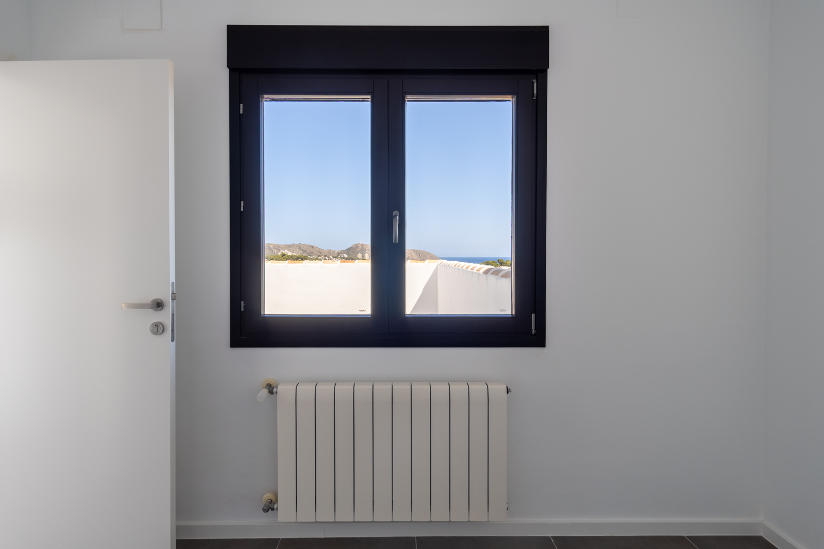 Dunkel gerahmtes Aluminiumfenster mit Doppelverglasung und Blick auf das Meer und Cabo de Moraira, platziert über einem weißen Heizkörper in einem hellen, modernen Raum.