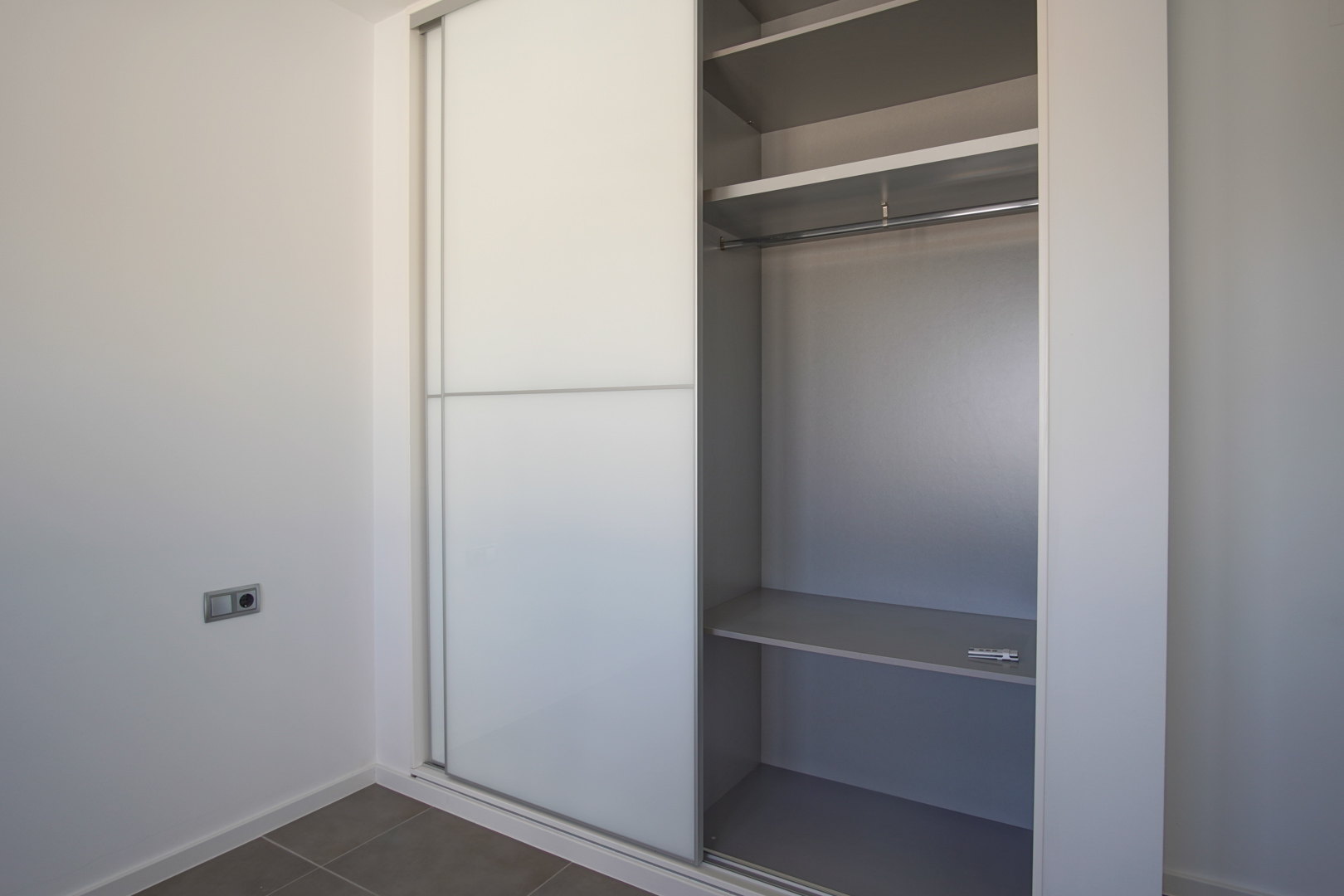 Einbauschrank mit weißen Schiebetüren und grauem Innenleben, ausgestattet mit Regalböden und Kleiderstange in einem modernen, schlichten Raum.